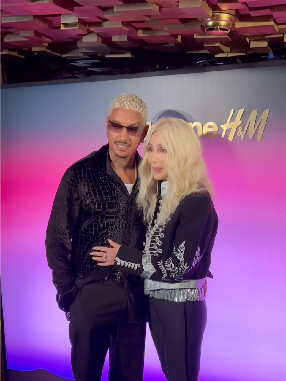 Cher and her boyfriend Alexander "A.E." Edwards standing together in front of an H&M backdrop, with pink and purple lighting in the background.