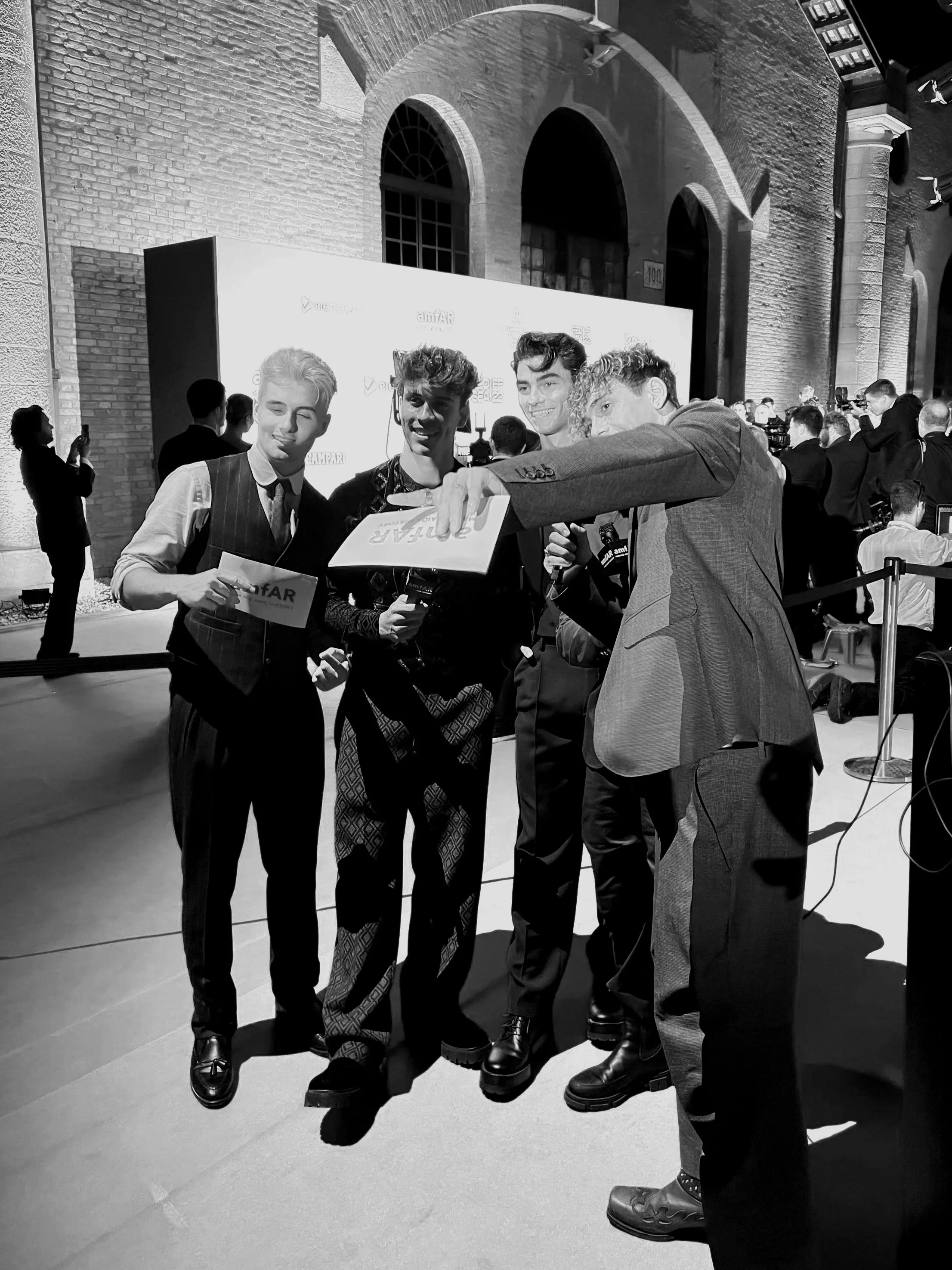 The Elevator Boys dressed in fashionable clothing are gathered together, smiling for a photo at an amfAR event with a crowd and a large screen in the background.