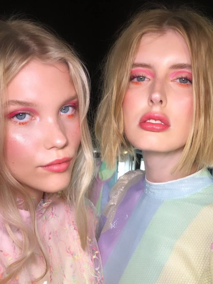 Two young women with blonde hair and pink makeup, wearing pastel-colored tops, posing for a selfie indoors.