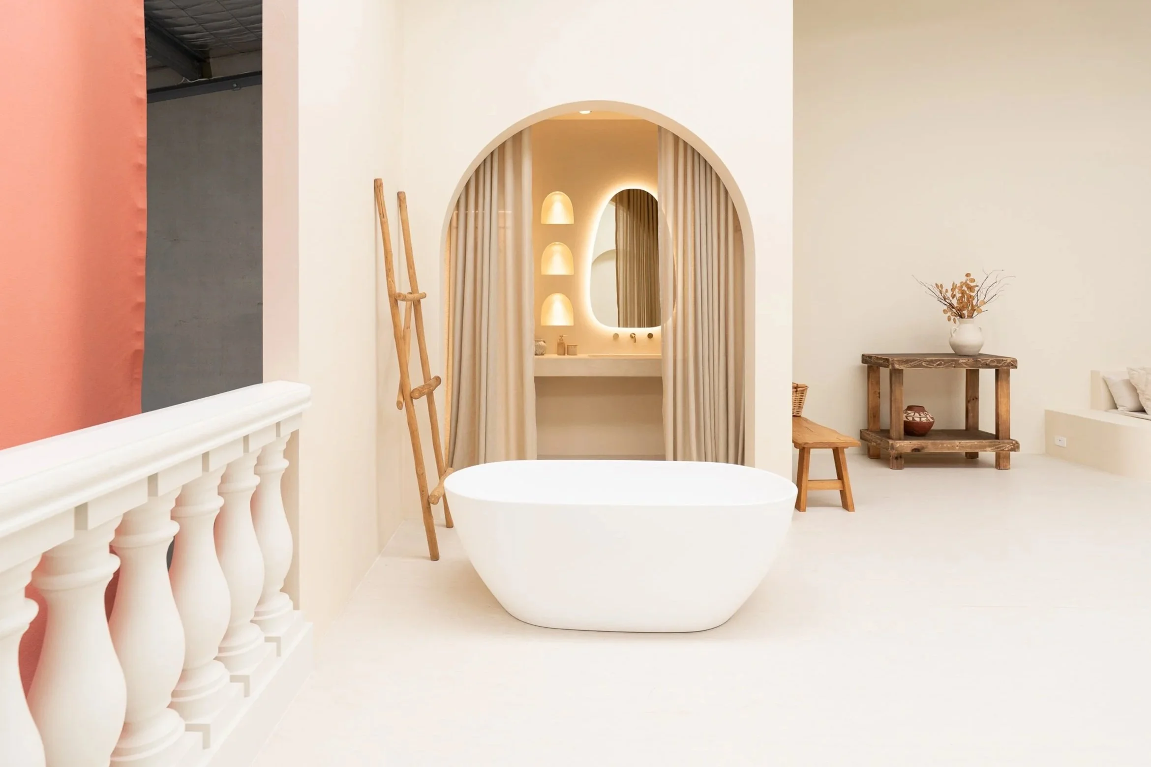 A cozy interior space with a wooden chair featuring cream cushions, large green indoor plants, a tall decorative mirror with a wavy border, and a beige wall with a vase of dried flowers in the background.