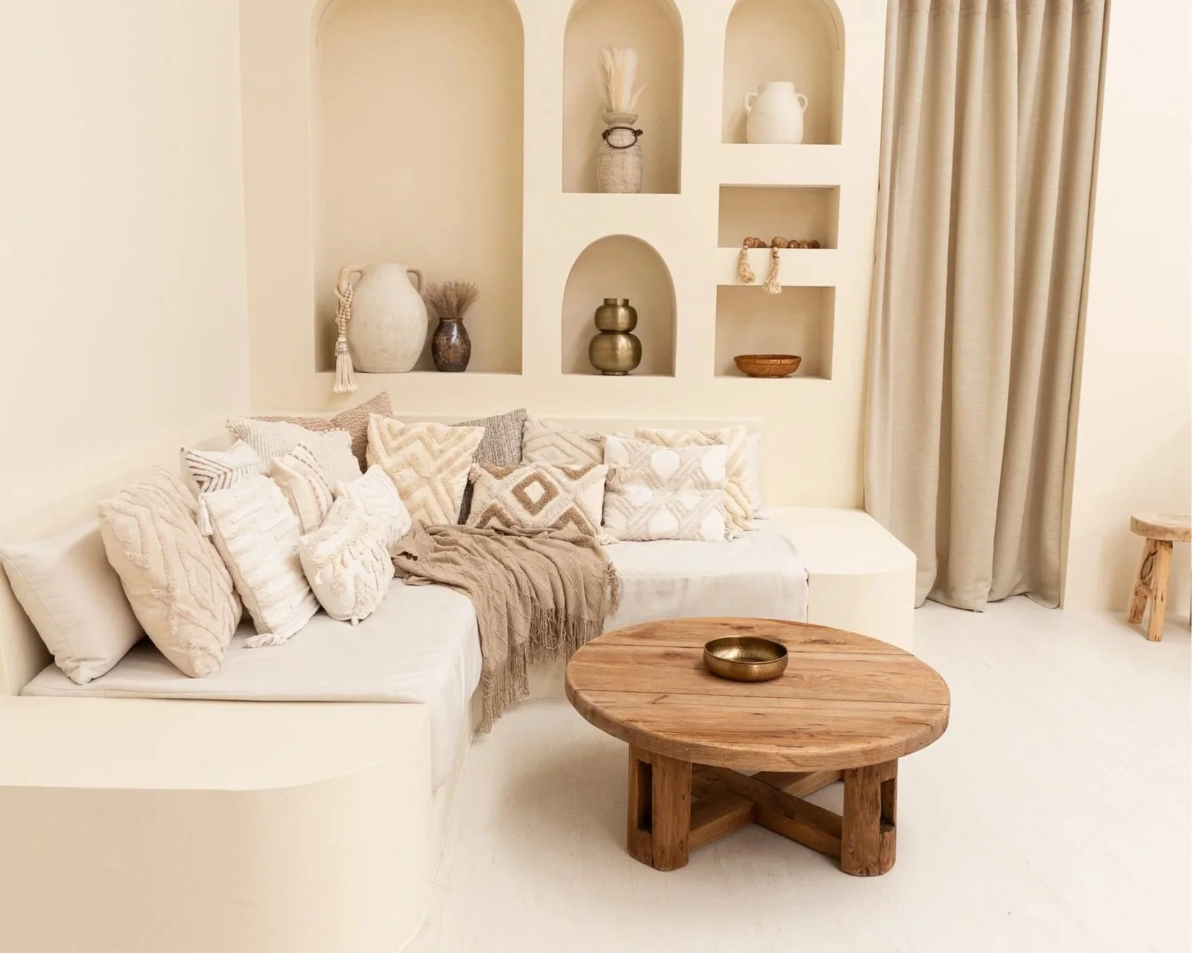 A simple interior corner with a wooden armchair that has white cushions, a small wooden side table, a large leafy plant behind the chair, a decorative mirror on the wall, and a small niche with a vase of dried flowers.