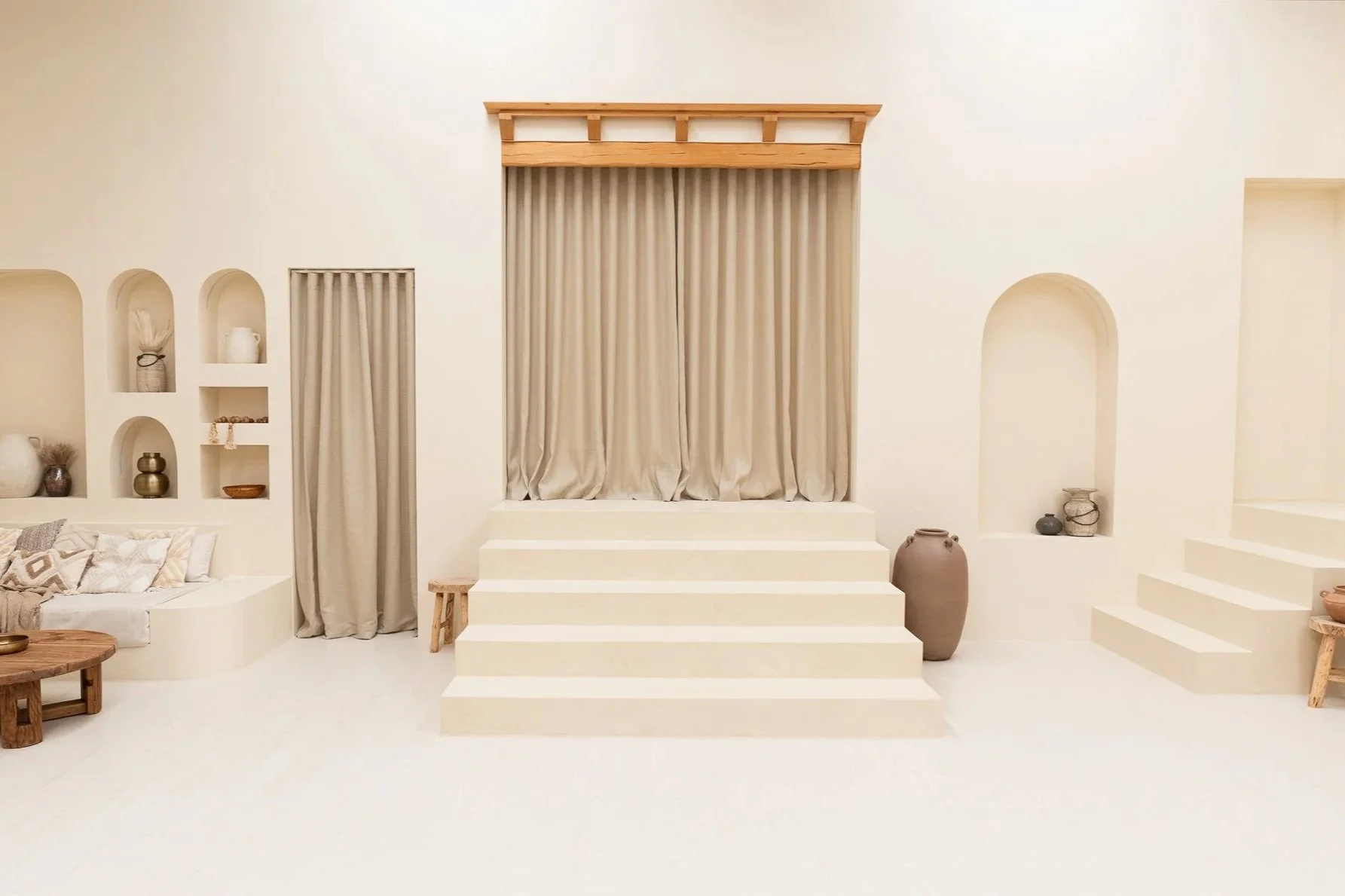 A room with white curtains, a decorative mirror with wavy edges, a potted plant with large green leaves, a wooden stool, and a curtain reflected in the mirror.