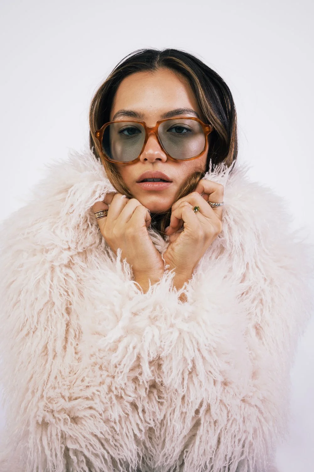 A woman wearing large sunglasses and a fluffy pink coat, holding her collar with both hands, against a plain white background.