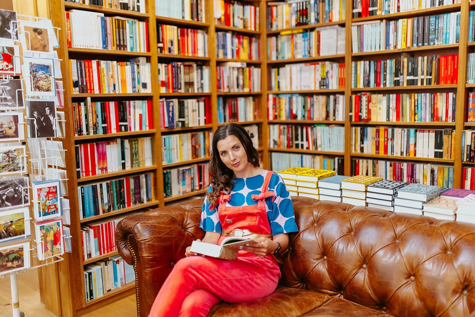 A woman sitting on a leather couch in a bookstore or library, holding a book. She has long dark hair and is wearing a blue and white polka dot top and red overalls. Behind her are tall wooden bookshelves filled with numerous books and a display rack with postcards or photographs.