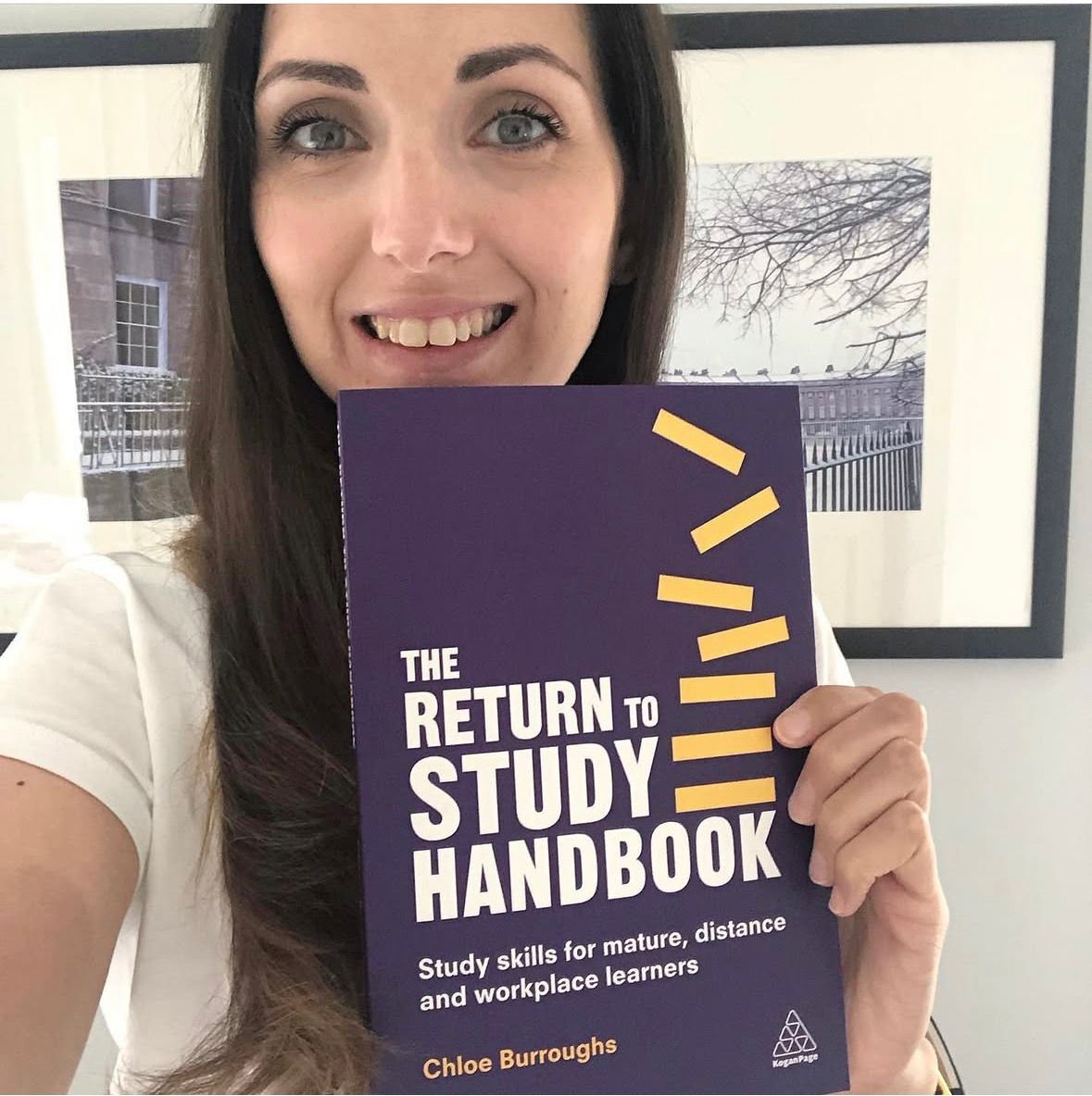 A woman holding a study guide titled 'The Return to Study Handbook' in front of her, with framed artwork of a winter scene with snow and bare trees behind her.