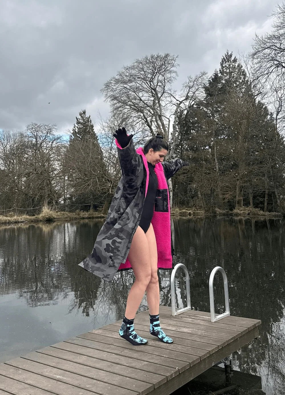 Chloe standing on a wooden dock by a lake, wearing a black swimsuit, a camouflage jacket, a pink scarf, and water shoes, with her arms raised and smiling. The background has leafless trees and cloudy skies.