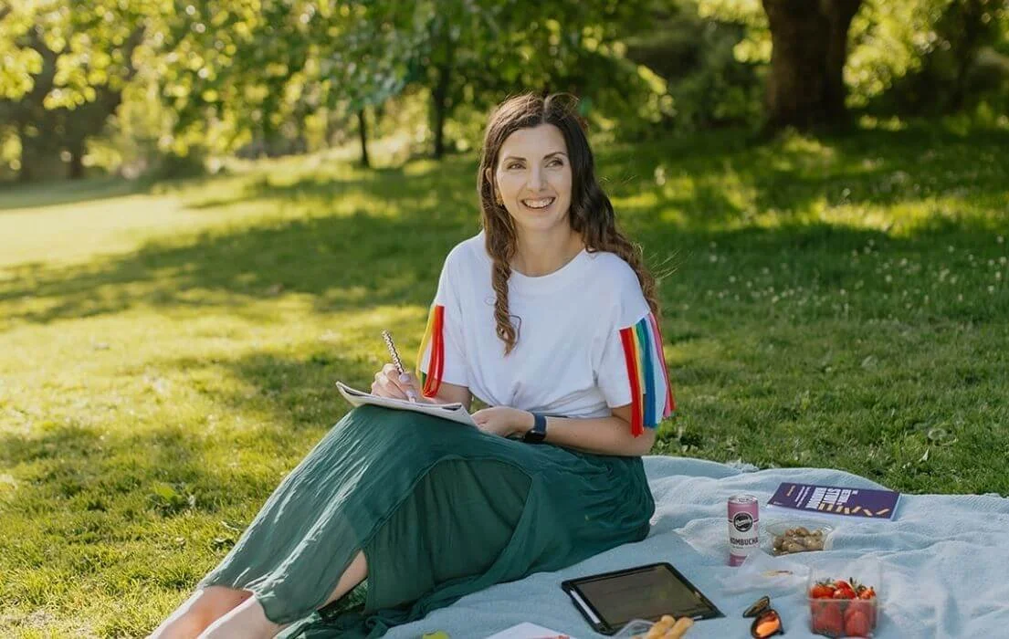 A woman sitting on a blanket in a park, smiling, with a notebook, tablet, and snacks around her.
