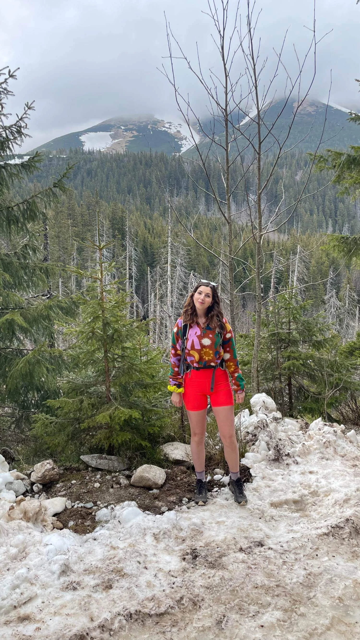 A woman standing on a snow-covered trail in a forest with mountain peaks in the background.