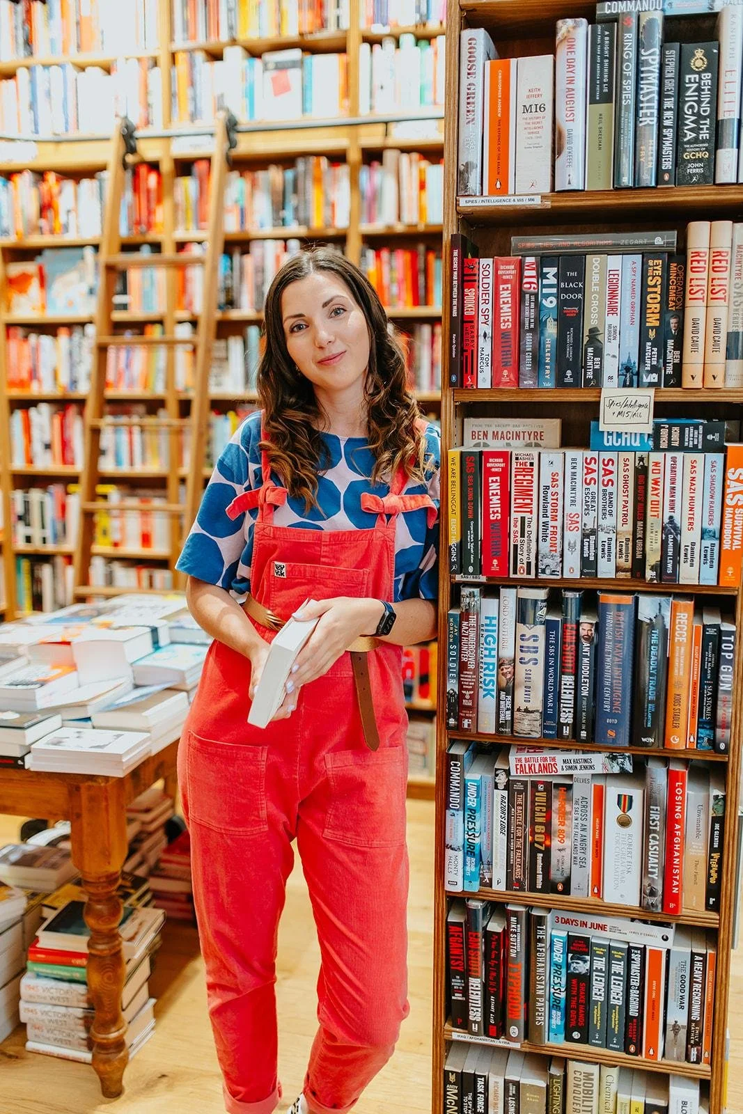 A young woman with long brown hair holding a book in a bookstore. She is wearing a blue polka-dot shirt, red overalls, and a smartwatch. Behind her are shelves full of books, and to her right is a tall bookshelf with various titles.