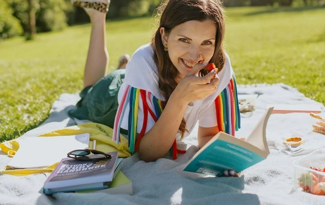 Chloe lying on her stomach on a picnic blanket in a park, smiling and eating a strawberry, with books, sunglasses, and snacks around her.