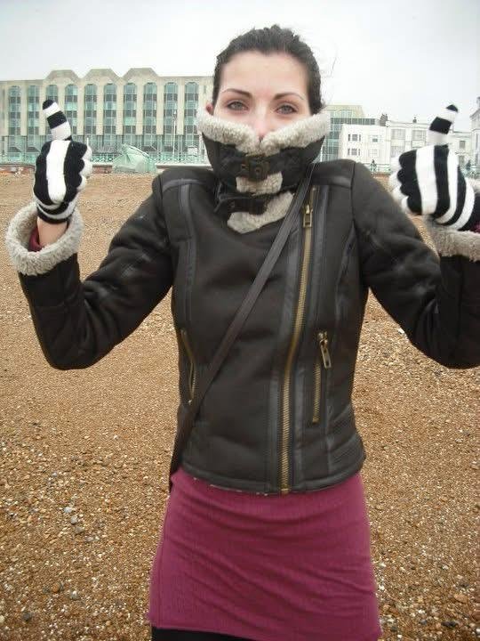 A woman wearing a black leather jacket, purple skirt, and striped gloves standing outdoors on a gravel surface, giving two thumbs up with a cityscape and cloudy sky in the background.