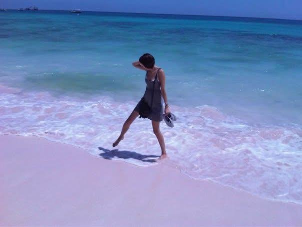 A woman wearing a gray dress and sunglasses, standing on a sandy beach near the water, holding her sandals, with the ocean and a boat in the background.