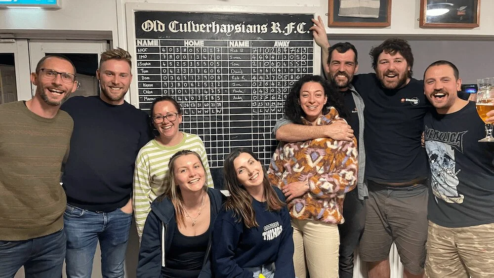 Group of nine friends smiling and celebrating in a pub, some hugging, standing in front of a board showing a golf score at Old Culverhaysians R.F.C.