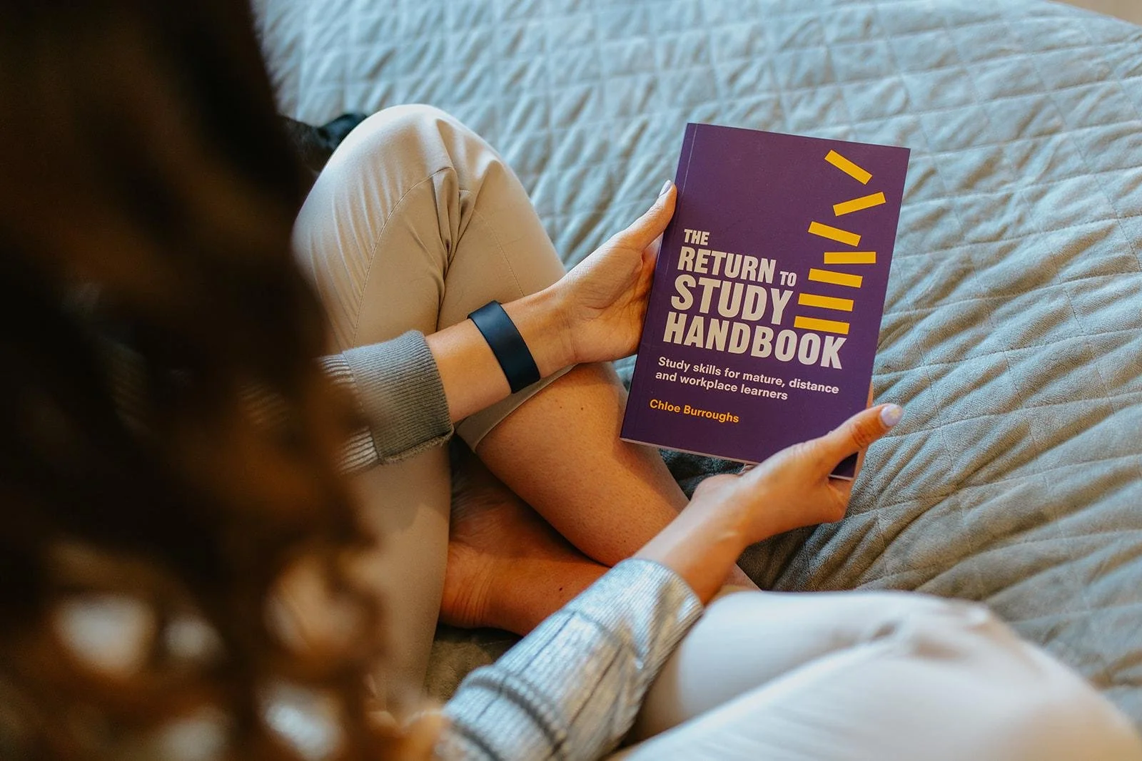 Chloe sitting on a quilted bedspread, holding a purple book titled 'The Return to Study Handbook.'