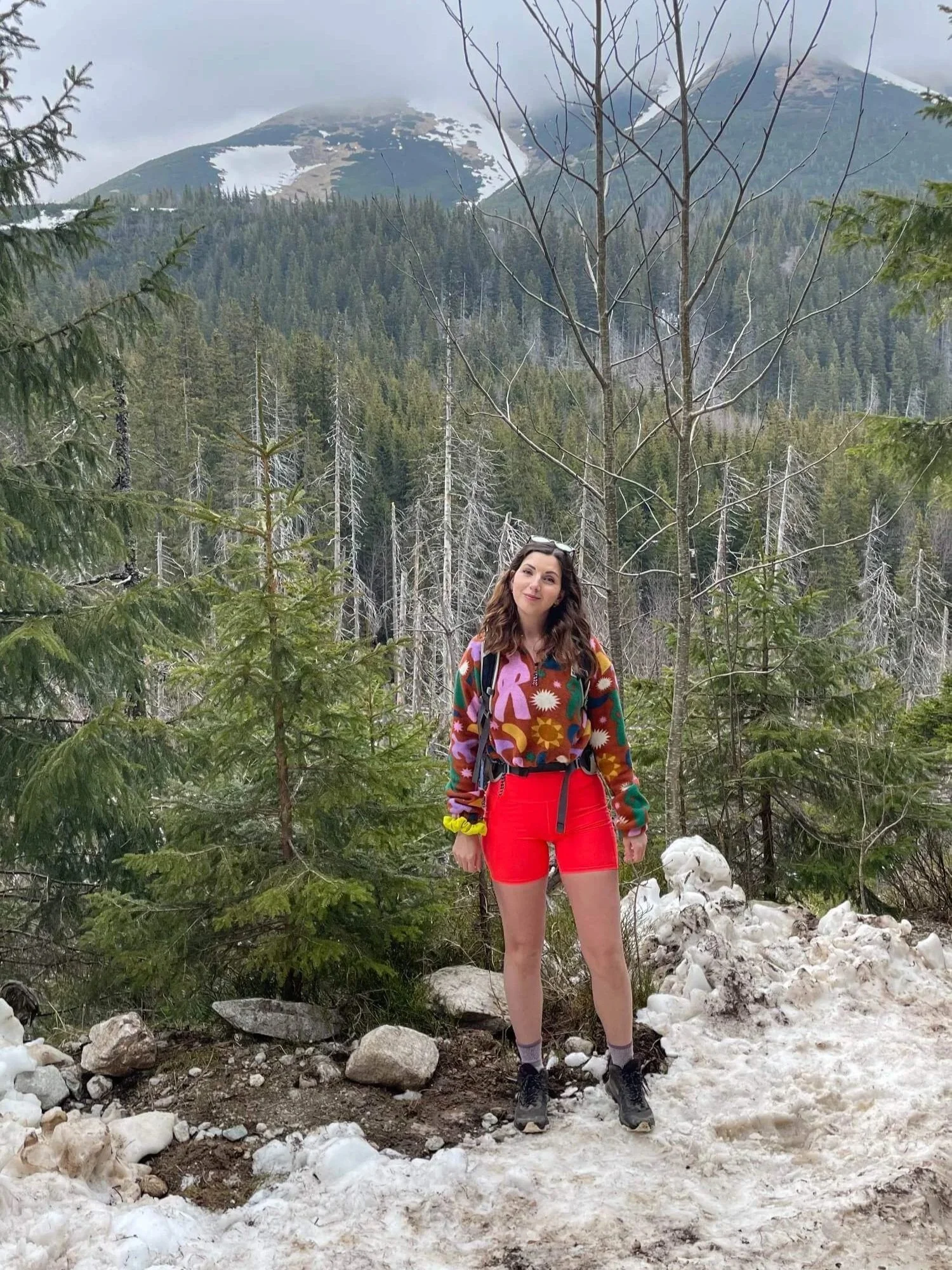 A woman with long brown hair standing in a mountain forest with snow, trees, and mountainous terrain in the background, wearing colorful clothing and hiking boots.