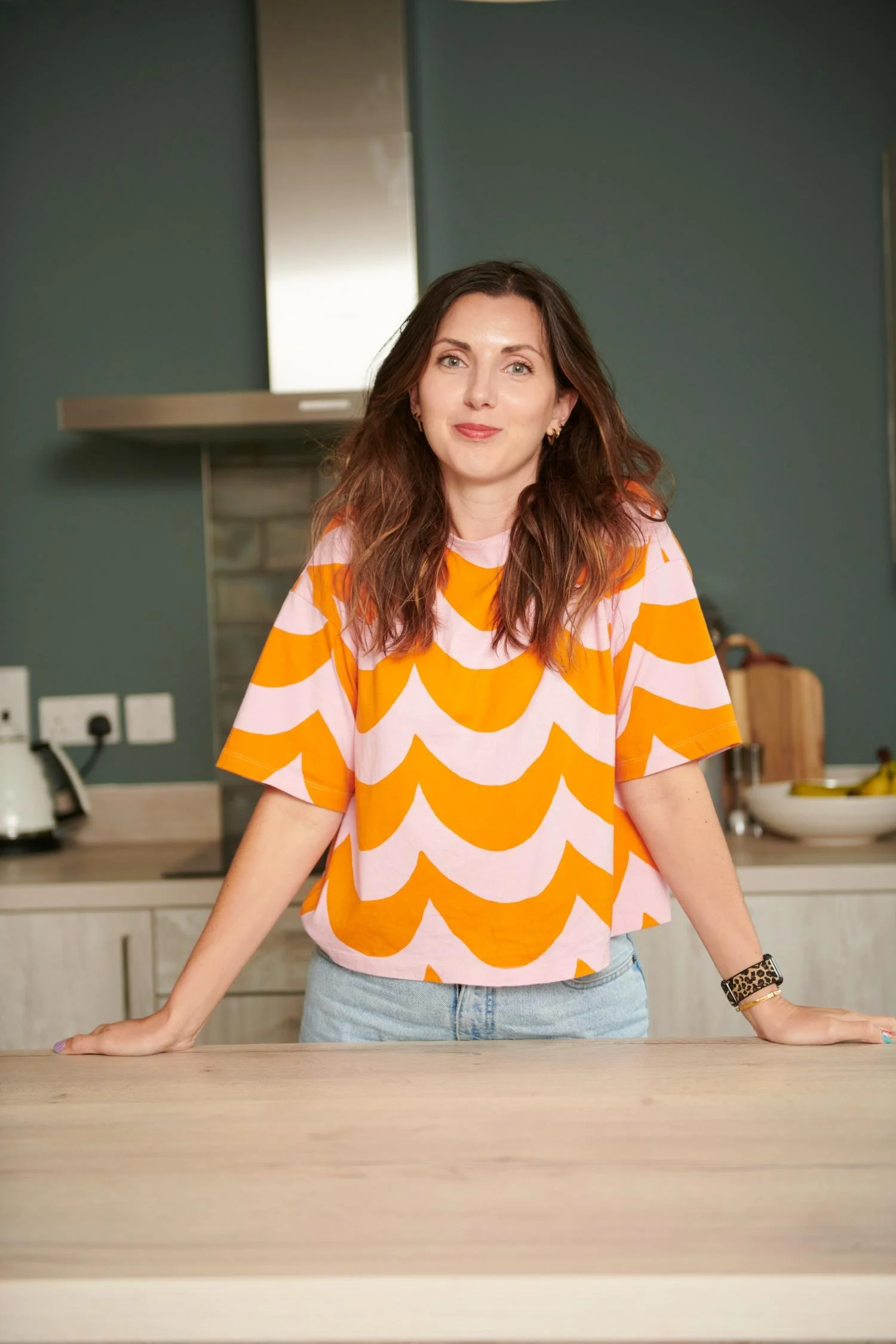 Chloe wearing a pink and orange patterned shirt, standing in a kitchen with teal walls and wooden countertop.