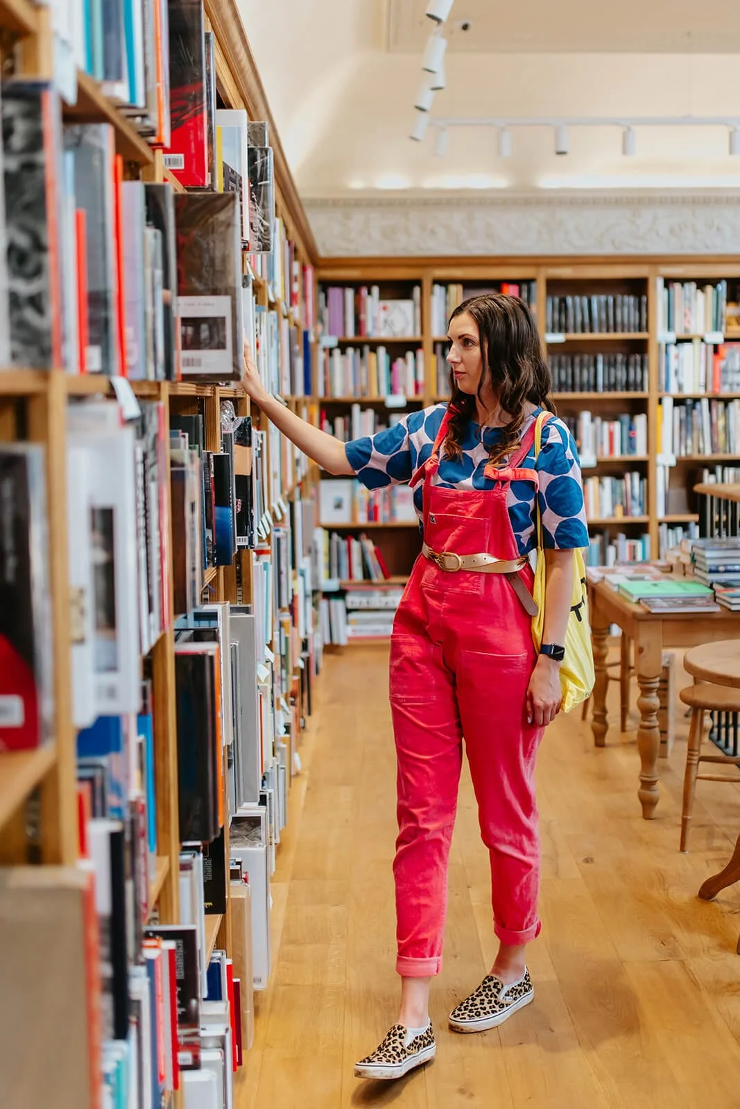 A woman with dark hair wearing a blue patterned shirt, red overalls, and leopard print slip-on shoes browsing books in a library.
