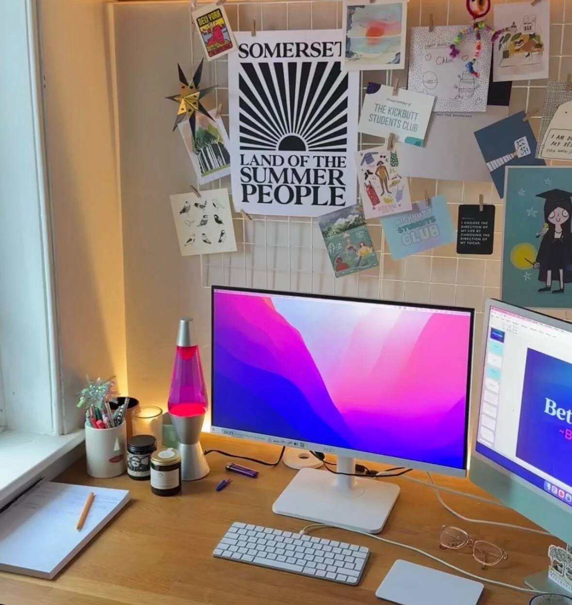 A home office desk with a dual monitor setup, a lava lamp, pens, notebooks, glasses, and decorative items. The wall behind features a white grid with various posters, artwork, and notes pinned, including a prominent black and white poster reading 'SOMERSET LAND OF THE SUMMER PEOPLE'.