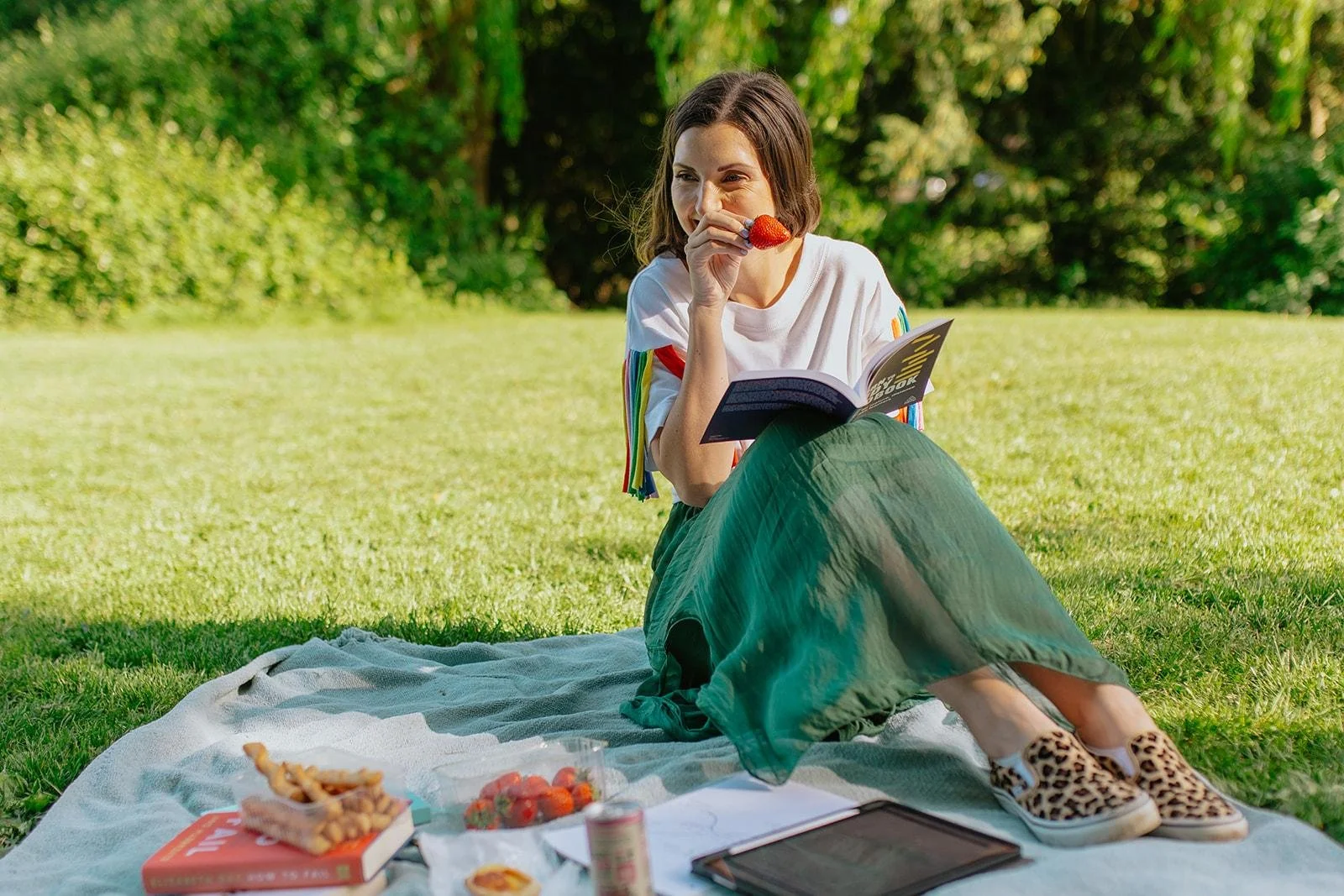 Chloe sitting on a picnic blanket in a park, eating strawberries, reading a book, next to snacks and beverages, wearing a white shirt, green skirt, and leopard print trainers.