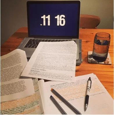 A laptop, open books with highlighted text, pens, and a glass of water on a wooden table.