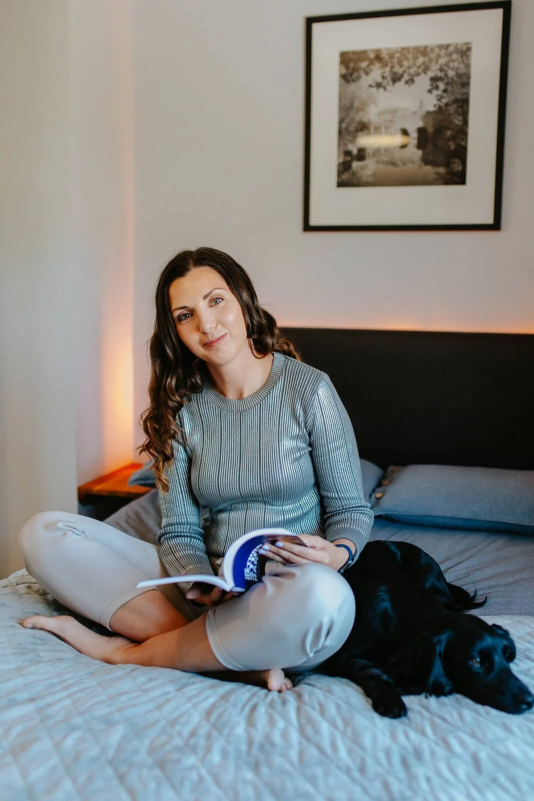Chloe sitting on a bed with gray bedding, smiling, holding a book, and a black dog lying beside her.
