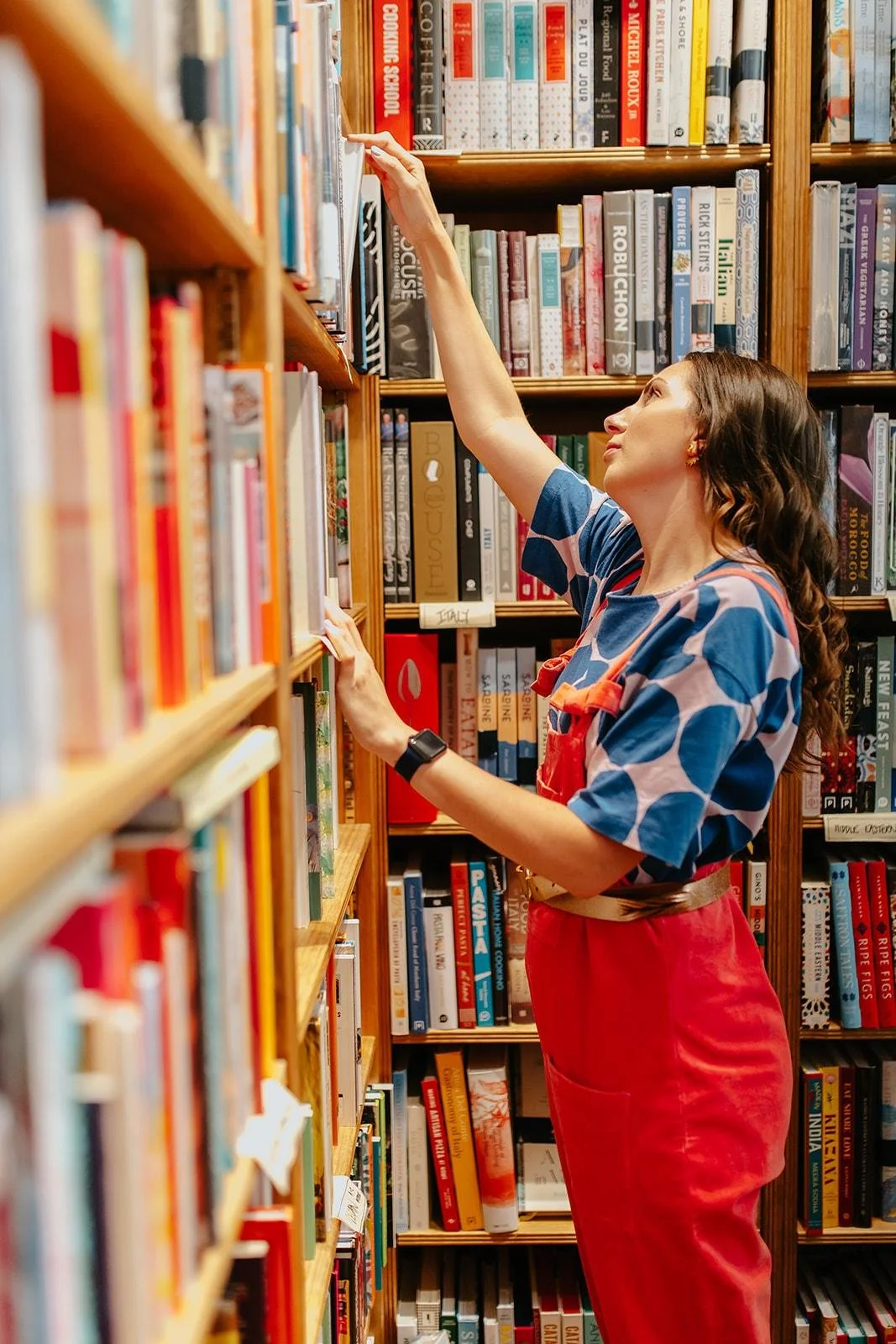 Chloe browsing books in a bookstore, reaching for a book on a tall wooden shelf.