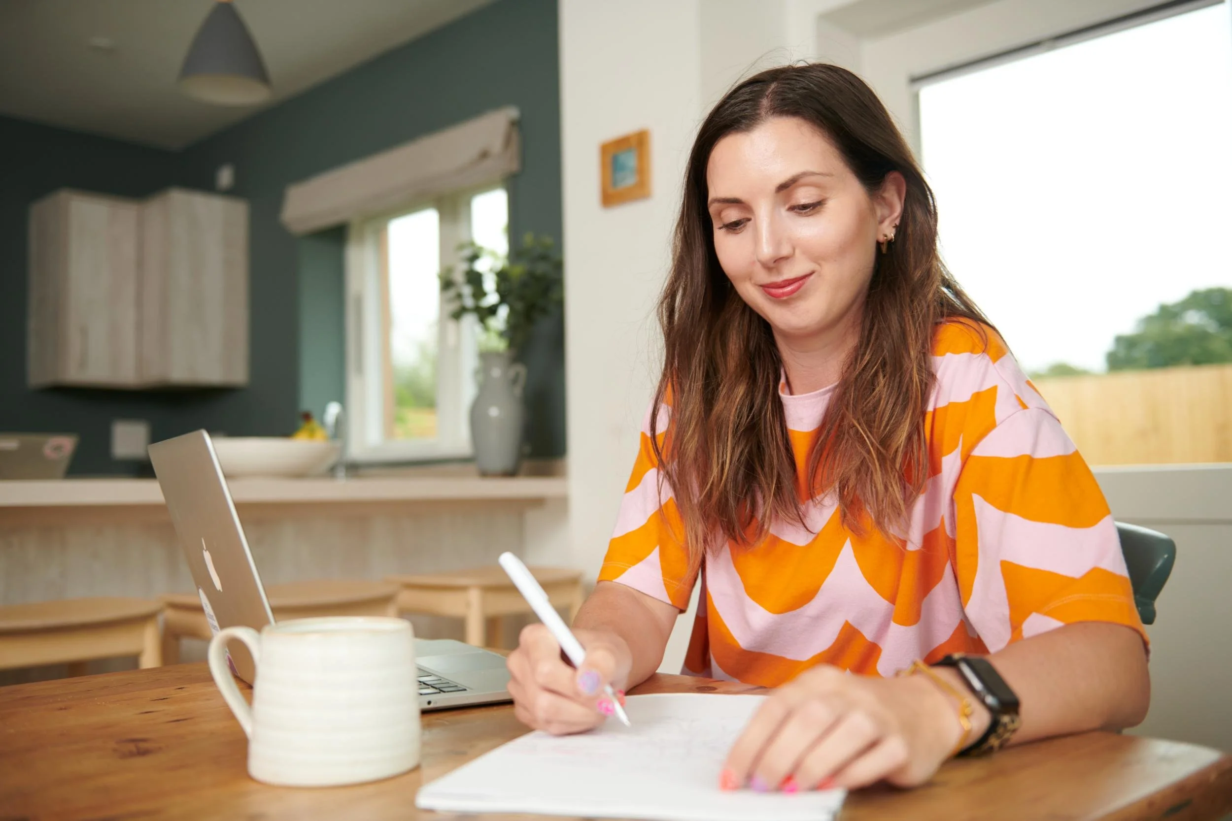A woman with long brown hair is sitting at a wooden table, writing in a notebook with a pen, in a bright kitchen. She is smiling and wearing a pink and orange patterned shirt, with a watch and earrings. There is a laptop, a white mug, and a beige ceramic cup on the table, and a window with a view of greenery in the background.