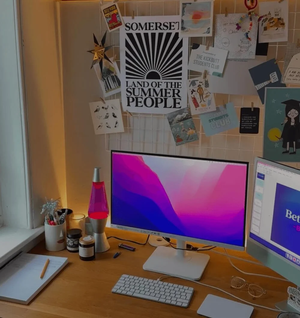 A work desk with two computer monitors, one displaying a pink and purple background, and the other showing a presentation slide. The desk has a notepad and pen, glasses, and a lava lamp. The wall behind is decorated with various posters, drawings, and cutouts, including a large black and white poster that reads "SOMERSET LAND OF THE SUMMER PEOPLE" and smaller notes and artwork pinned to a grid.