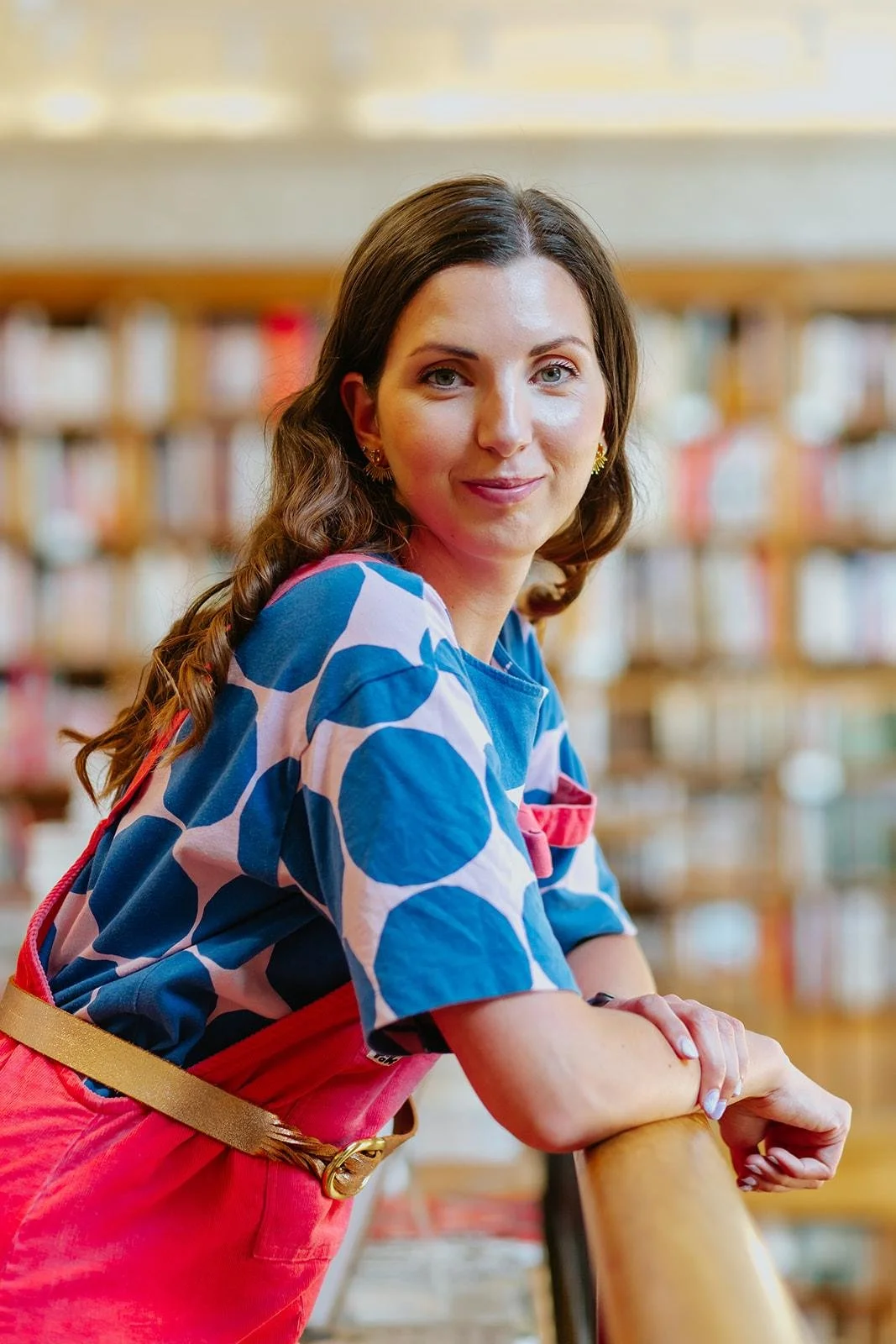 Chloe wearing a blue and white patterned top with red overalls, leaning on a wooden railing in a library, smiling at the camera with bookshelves in the background.