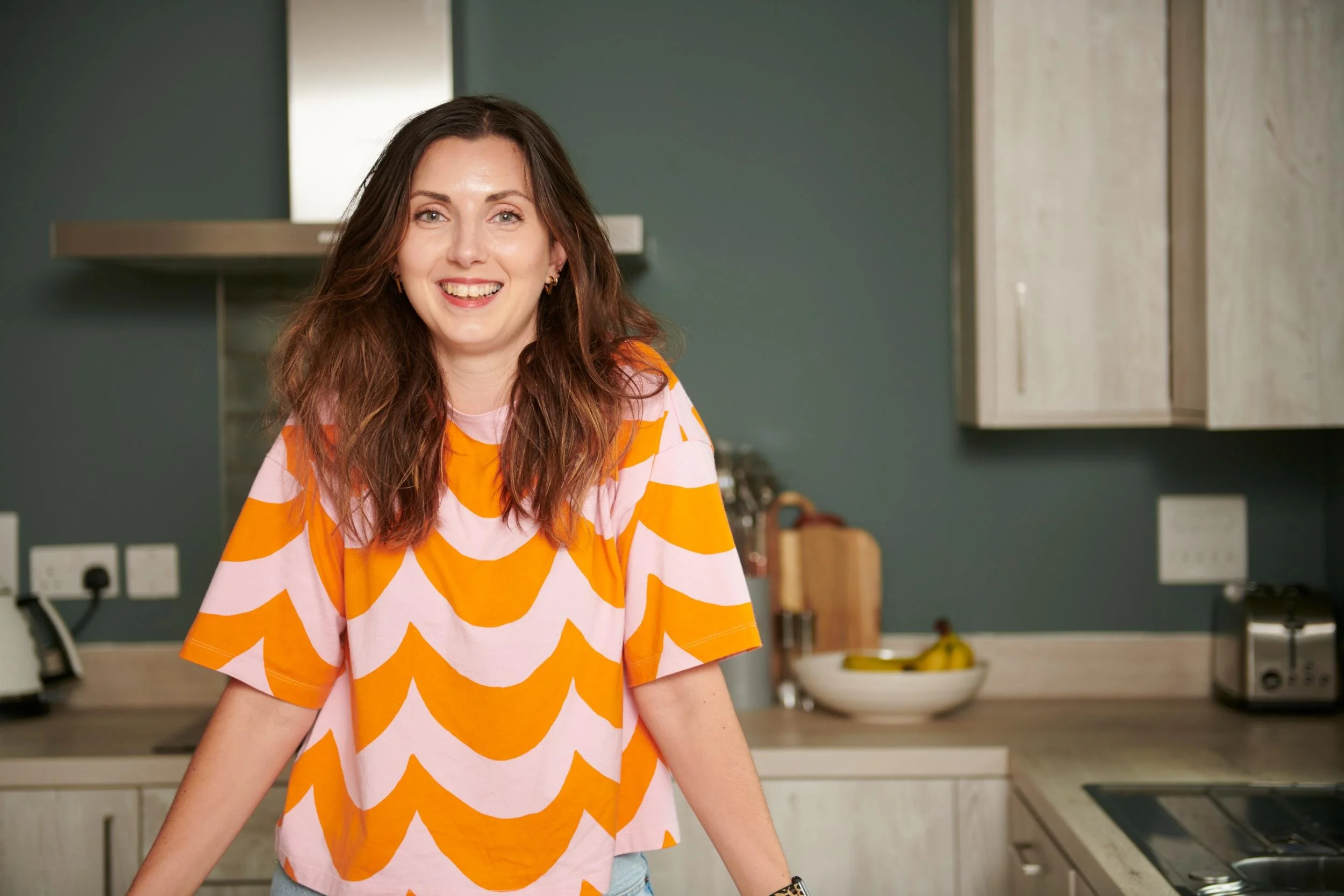 Close-up of a smiling woman with long brown hair wearing an orange and pink patterned t-shirt, standing in a modern kitchen.