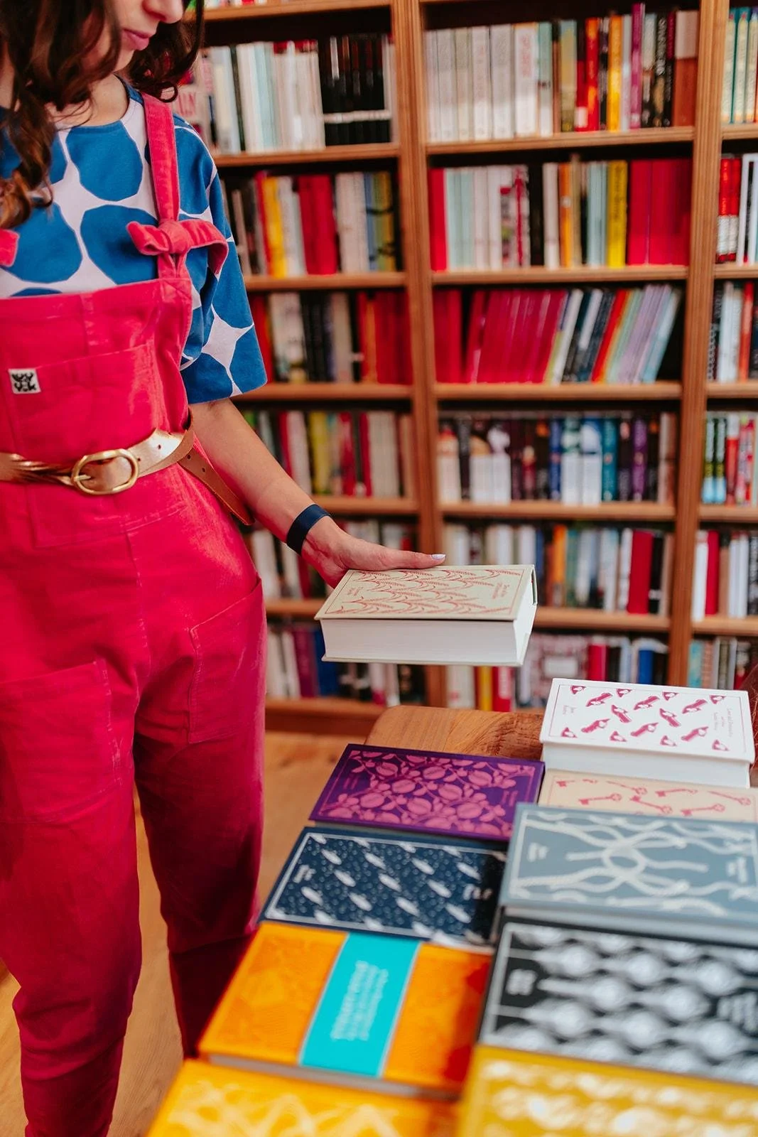 Chloe in vibrant red dungarees and a blue patterned shirt browsing books in a bookstore, with a wooden bookshelf filled with colourful books in the background, and books with colourful covers on a table in the foreground.