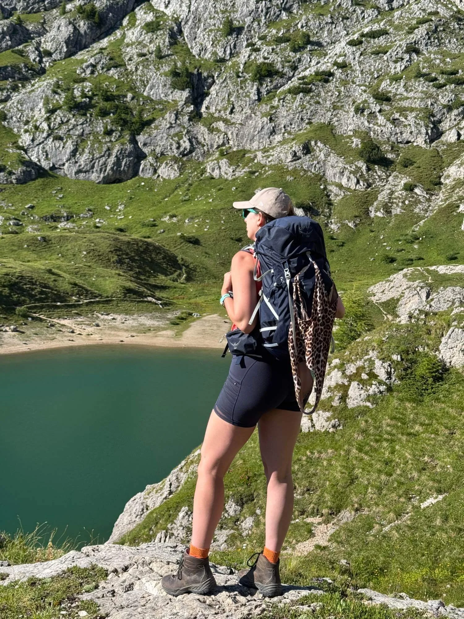 Chloe with a backpack looking at a green lake with rocky hills and mountains in the background during daytime.