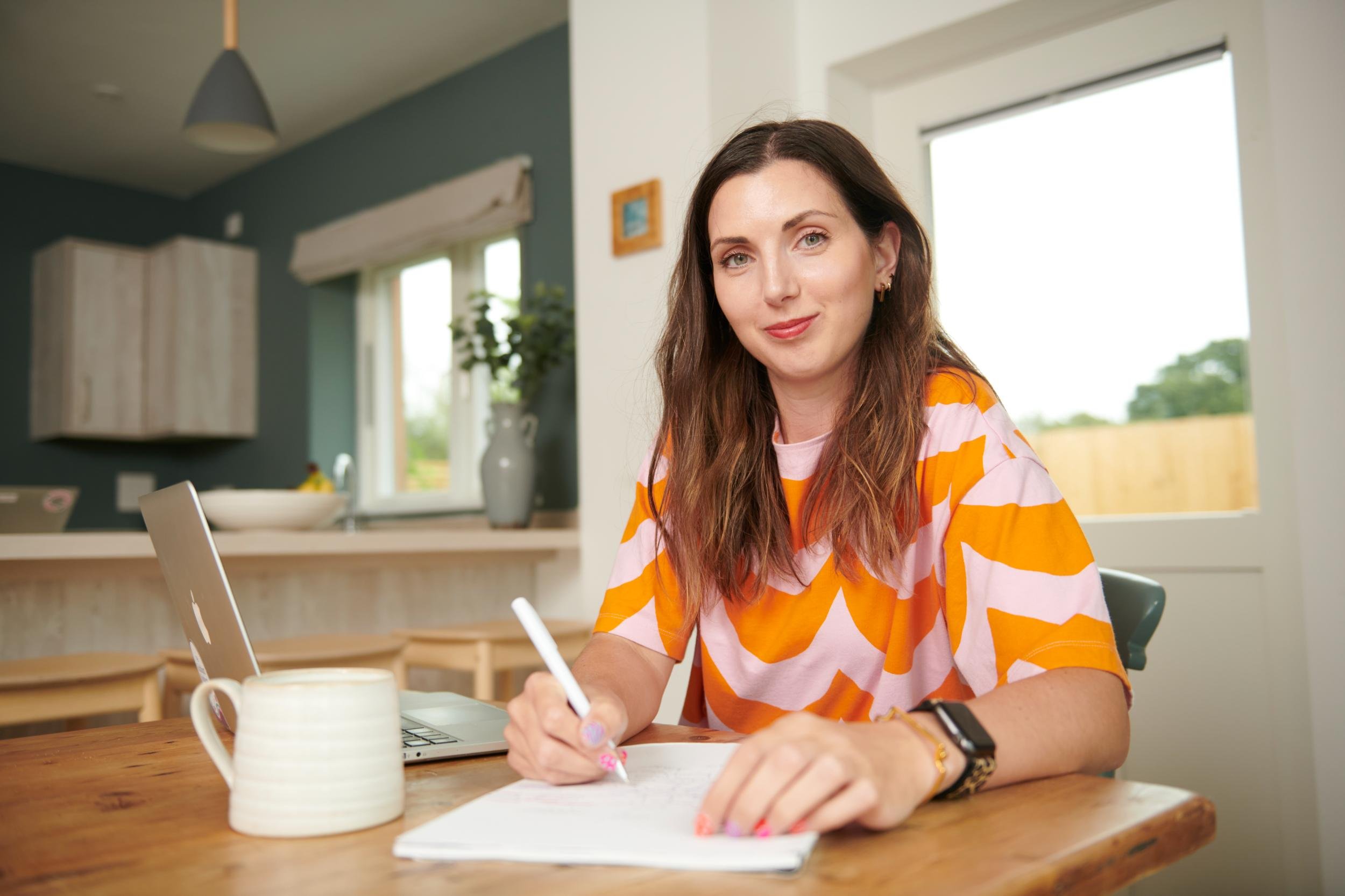 A woman with long brown hair wearing a pink and orange patterned T-shirt sitting at a wooden table, writing in a notebook with a pen. There is a white mug and a laptop in front of her in a bright, modern kitchen.
