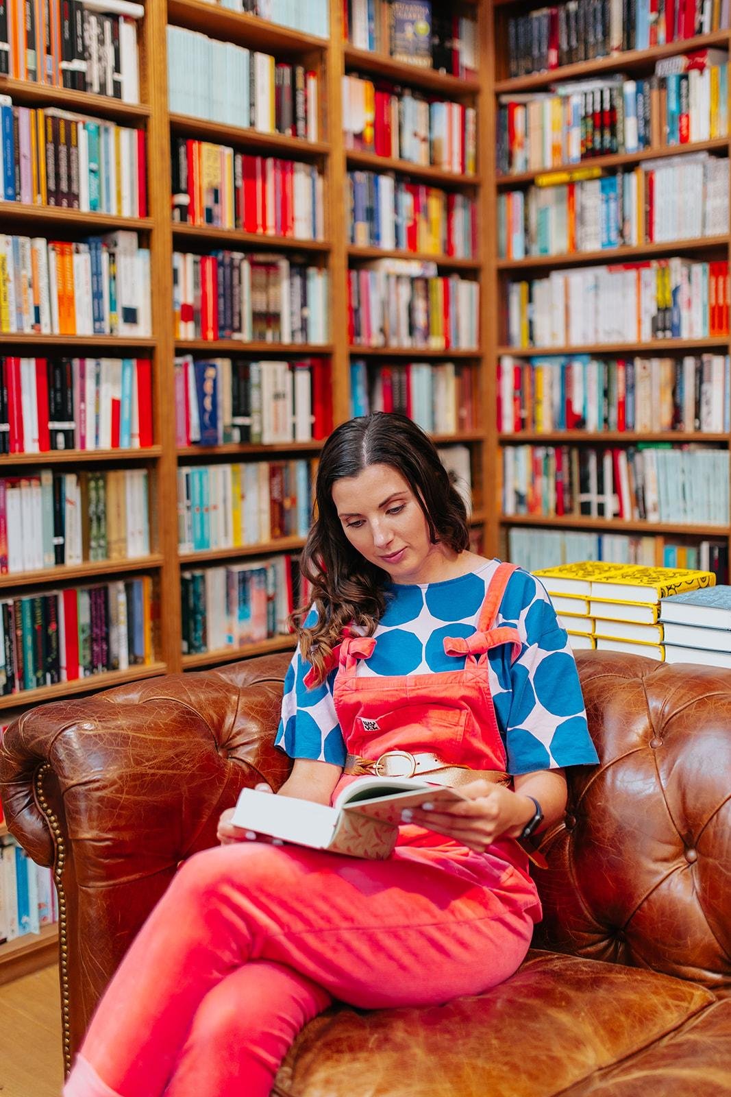 A woman with dark wavy hair in a blue and white patterned shirt and orange overalls sitting on a brown leather couch reading a book in a cozy library surrounded by shelves filled with books.