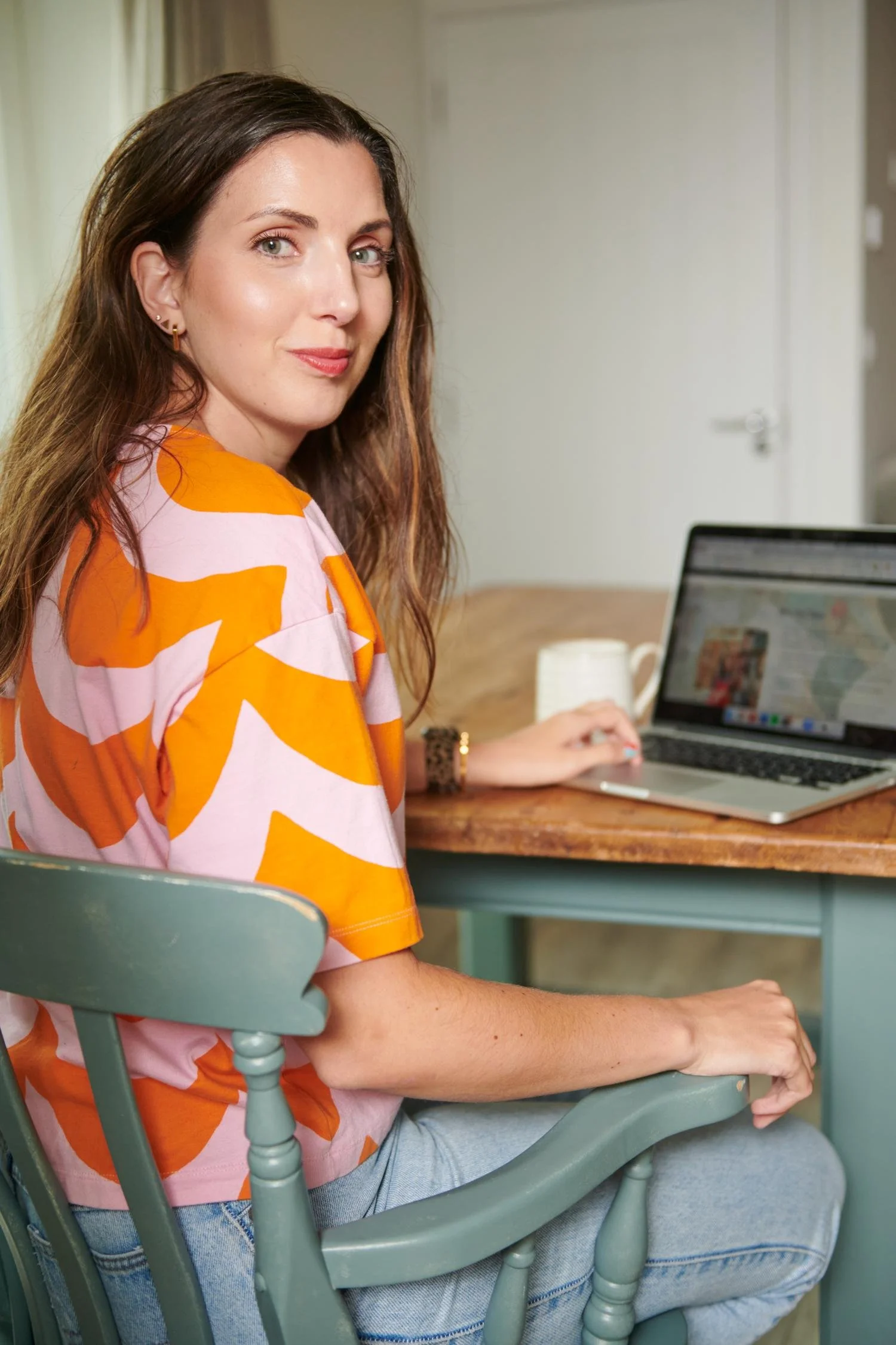A woman with long brown hair, wearing a pink and orange patterned top, sitting at a wooden table with a laptop and a white mug, in a room with light-colored walls and curtains.