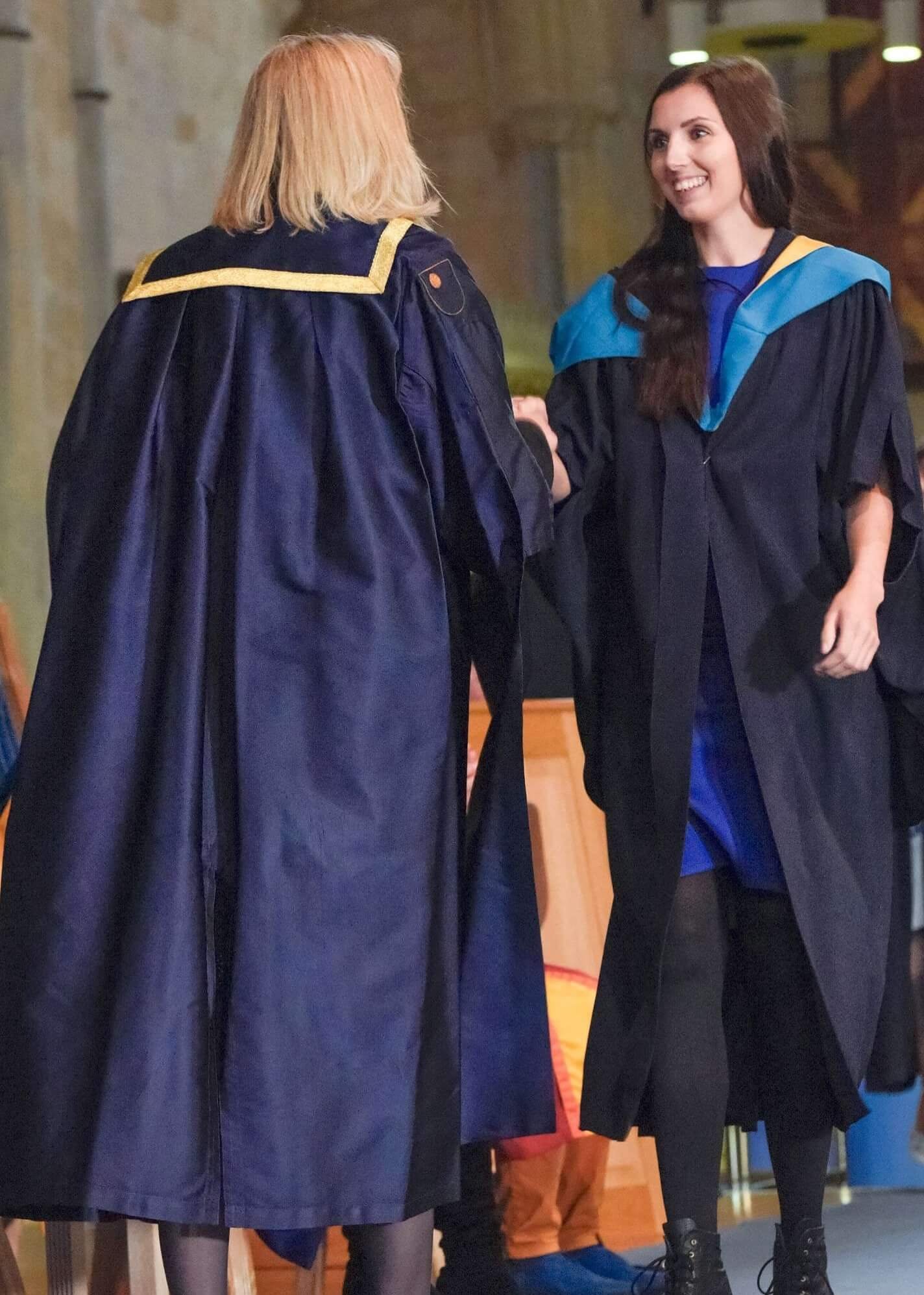 Two women in graduation gowns shaking hands during a ceremony. One woman has blonde hair and the other has long dark hair. The woman with dark hair is smiling.