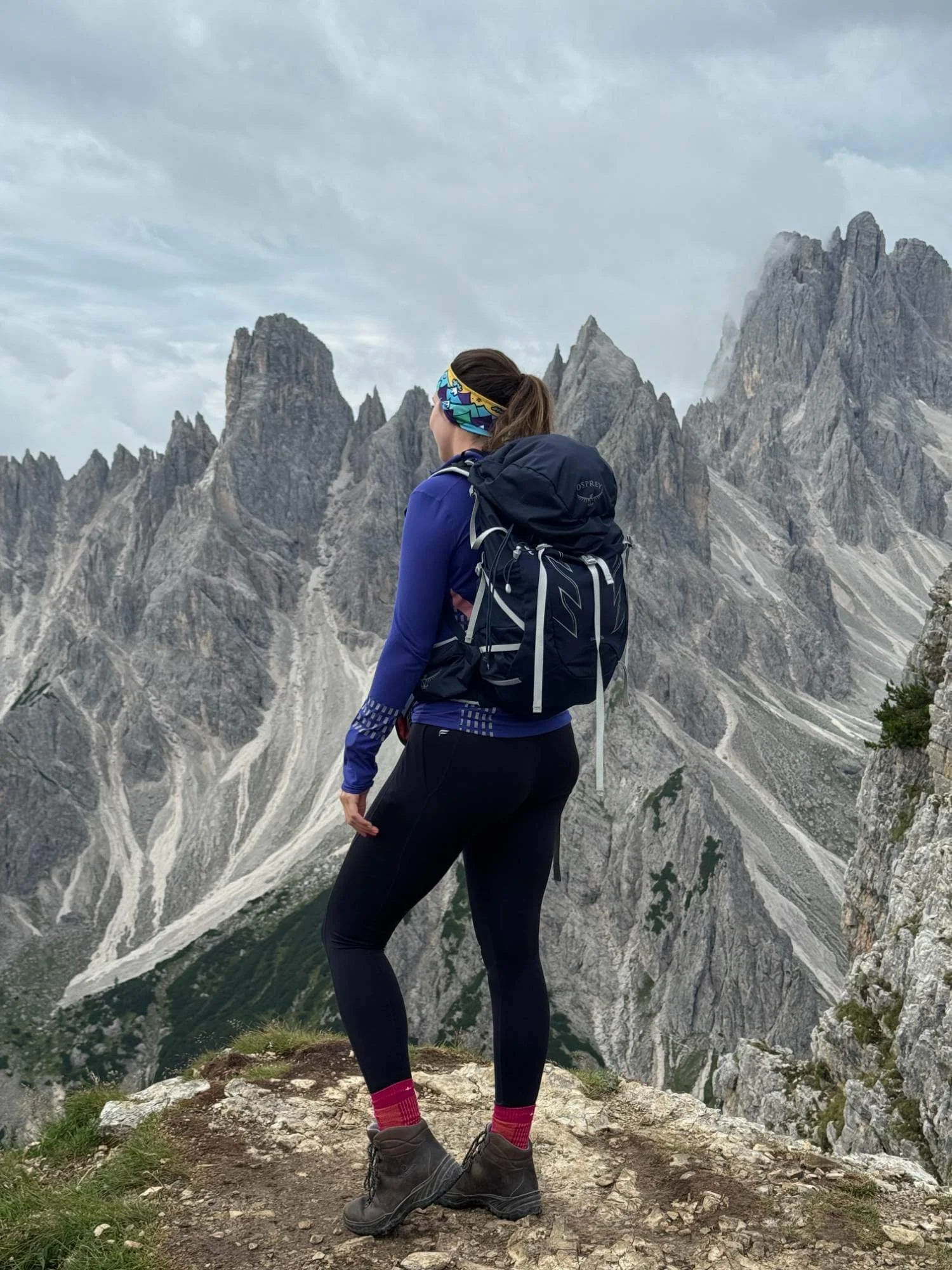 Chloe with a backpack and colourful headband standing on rocky terrain, overlooking jagged mountain peaks and a cloudy sky.