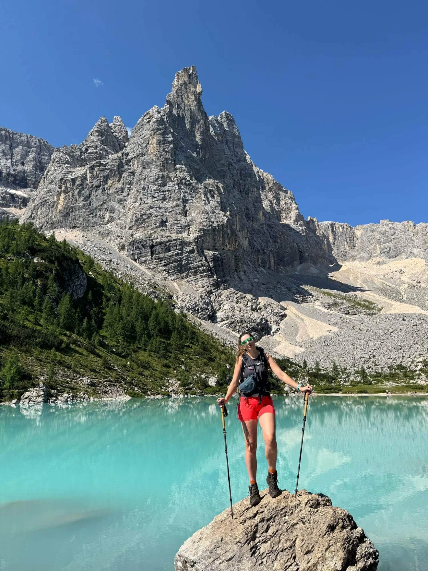 Chloe hiking near a turquoise lake in a mountainous area with tall rocky peaks and green trees.