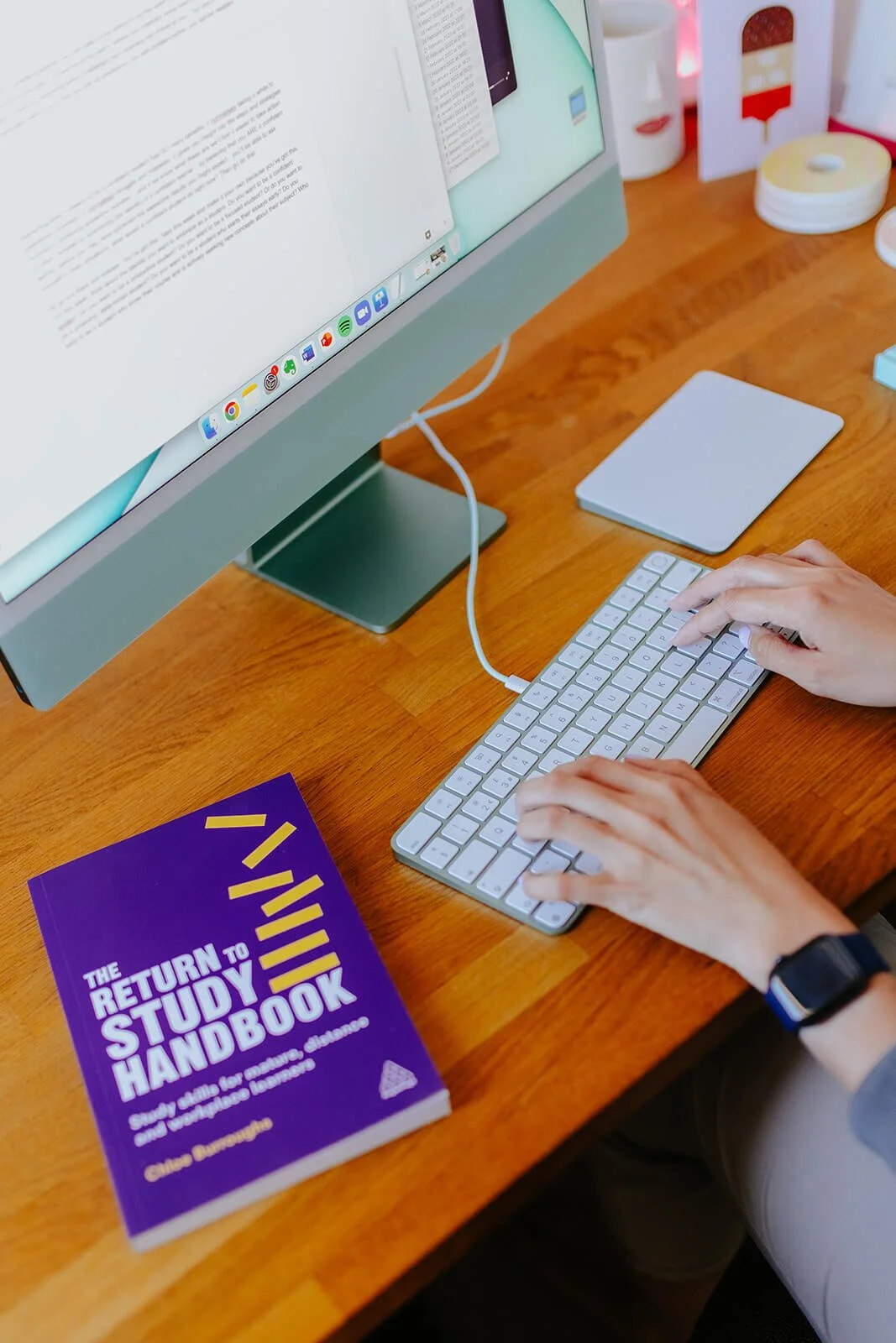 Chloe working on an iMac computer with a wireless keyboard at a wooden desk. There is a purple study guide titled "The Return to Study Handbook" on the desk, along with various office supplies and decorative items in the background.