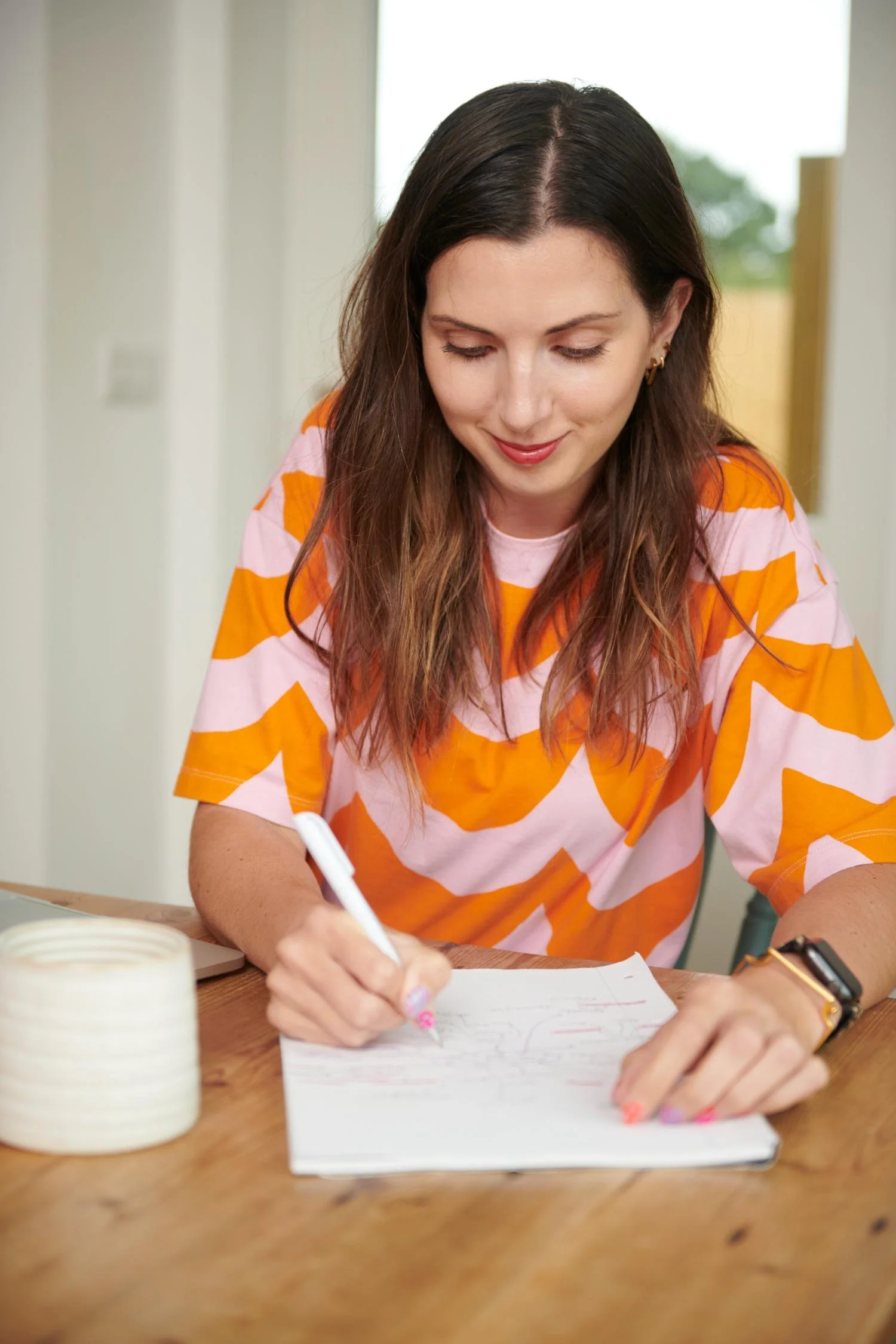A woman with long brown hair, wearing a pink and orange patterned shirt, is sitting at a wooden table writing in a notebook with a white pen. There is a roll of masking tape on the left side of the table, and a smartwatch on her left wrist. The background is indoors with a window showing greenery outside.