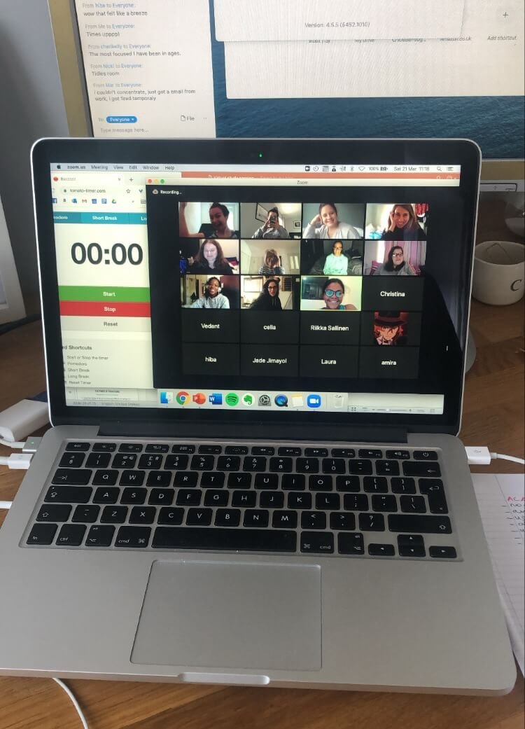 A laptop on a wooden desk displaying a Zoom video conference with multiple participants. Behind it, a larger computer monitor shows a document or webpage. To the right, there is a white mug and some papers.