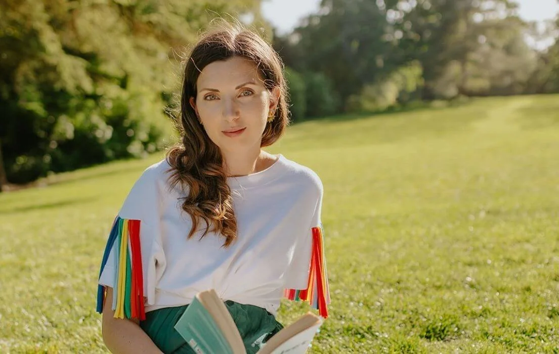 Young woman sitting on grass in park, holding a book, with trees and sunlight in background.