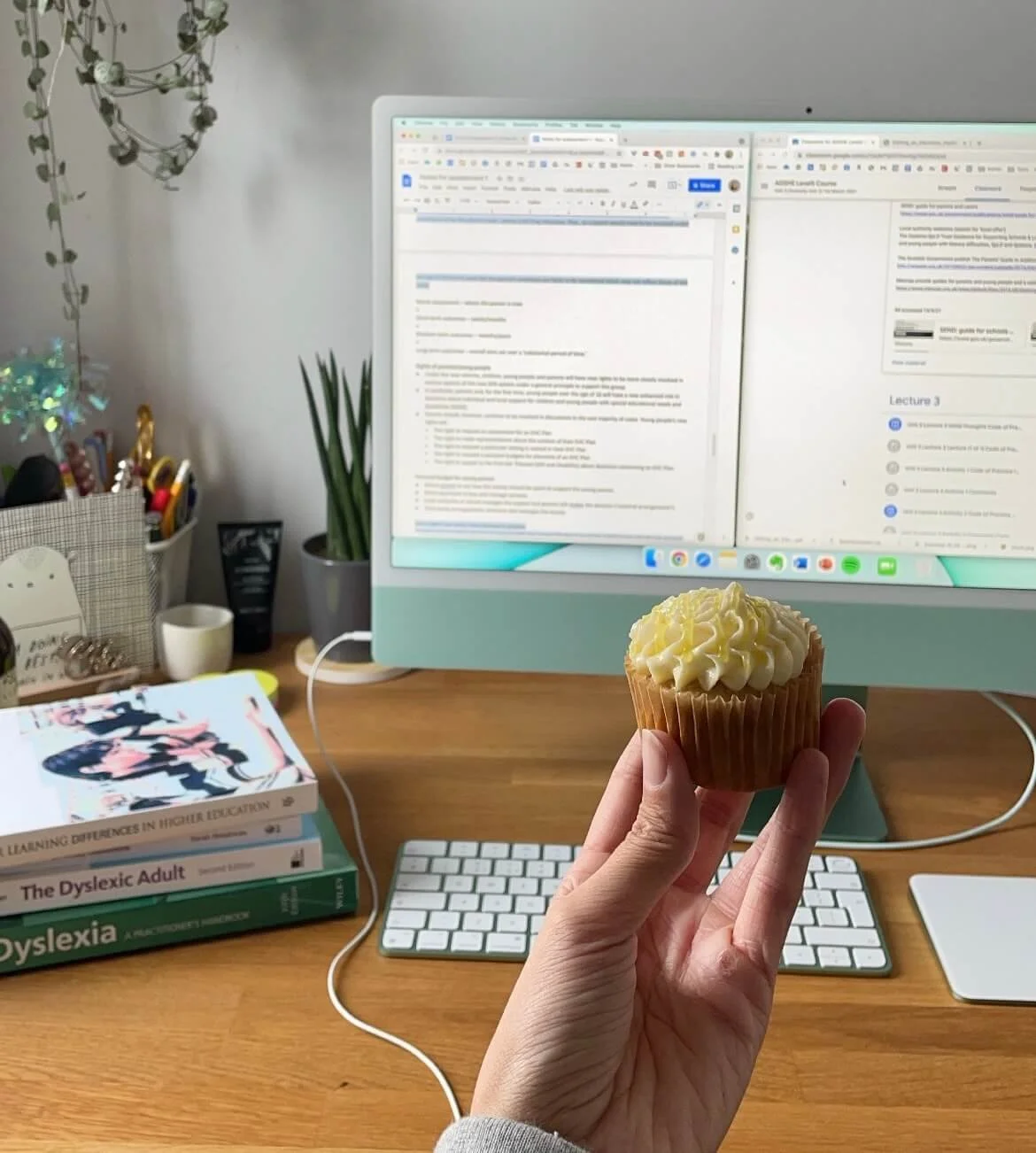 A person holding a cupcake with white frosting in front of a computer workspace with books, a keyboard, and a monitor displaying documents.
