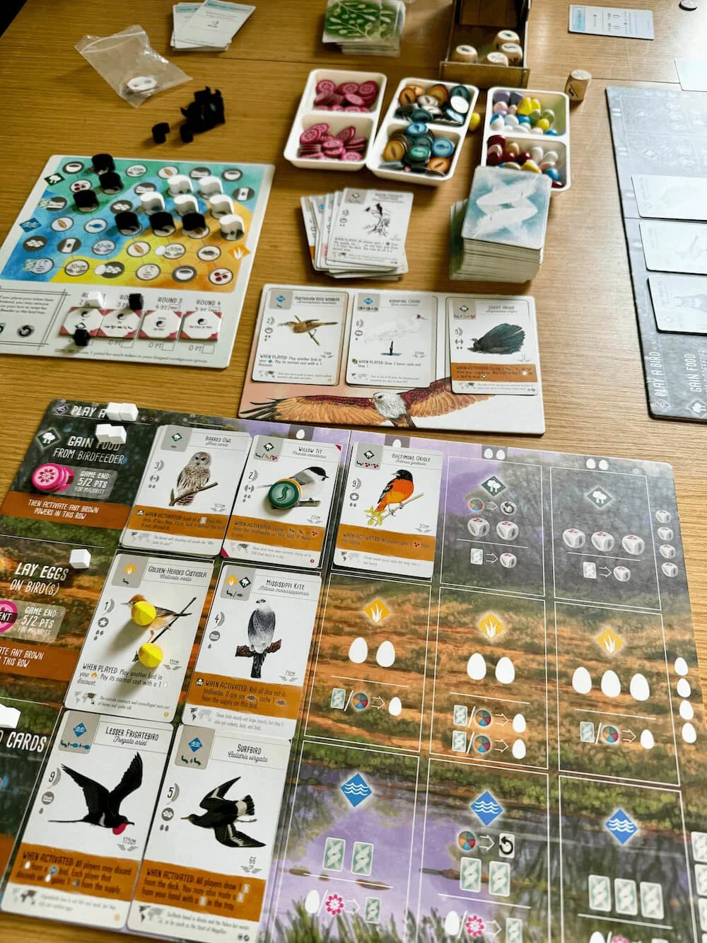Board game setup with various cards, tokens, and game pieces on a wooden table.