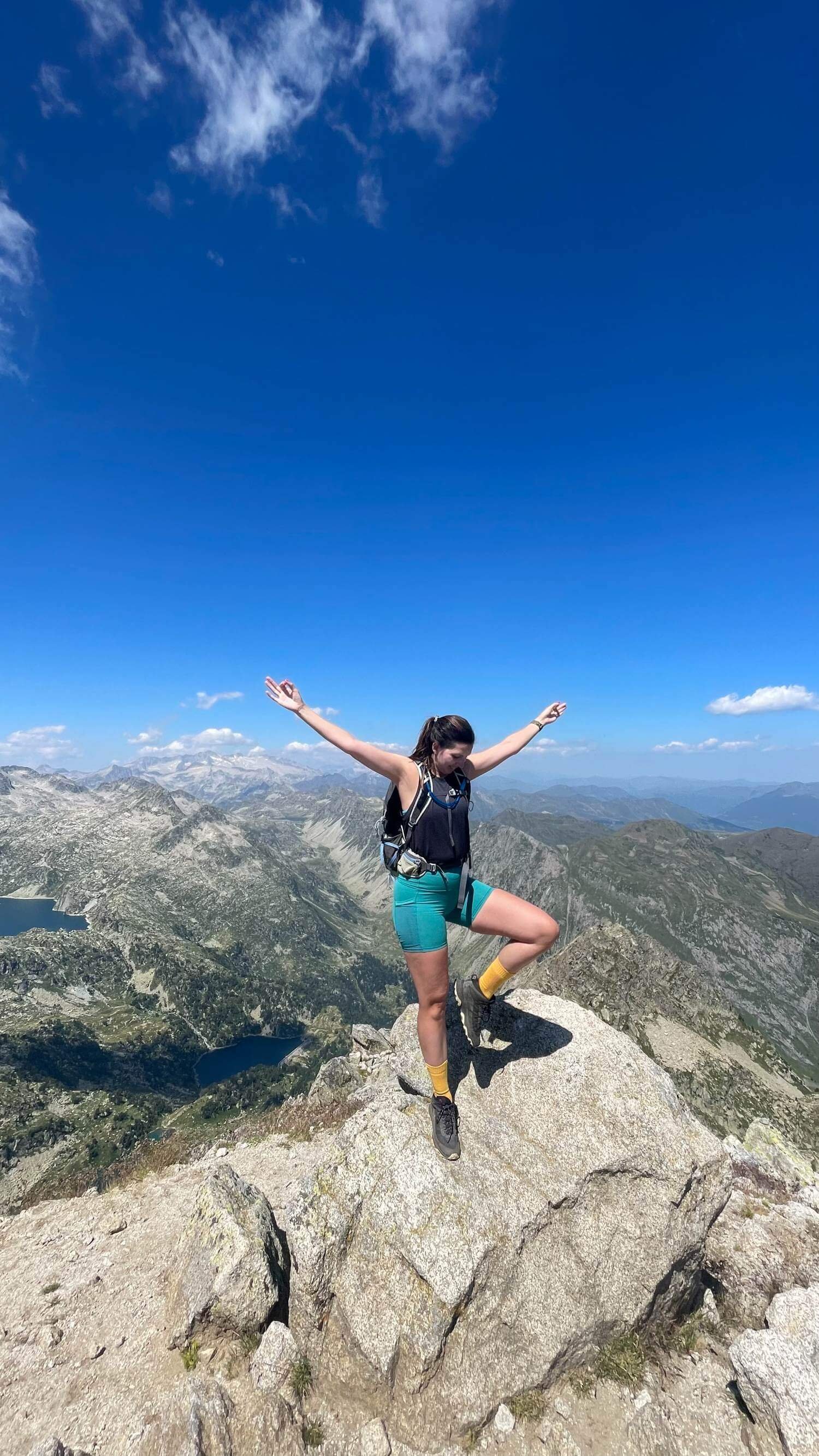 Chloe with a backpack standing on a rock with mountains and a lake in the background, arms raised in the air, enjoying the view from a mountain summit under a bright blue sky with a few clouds.