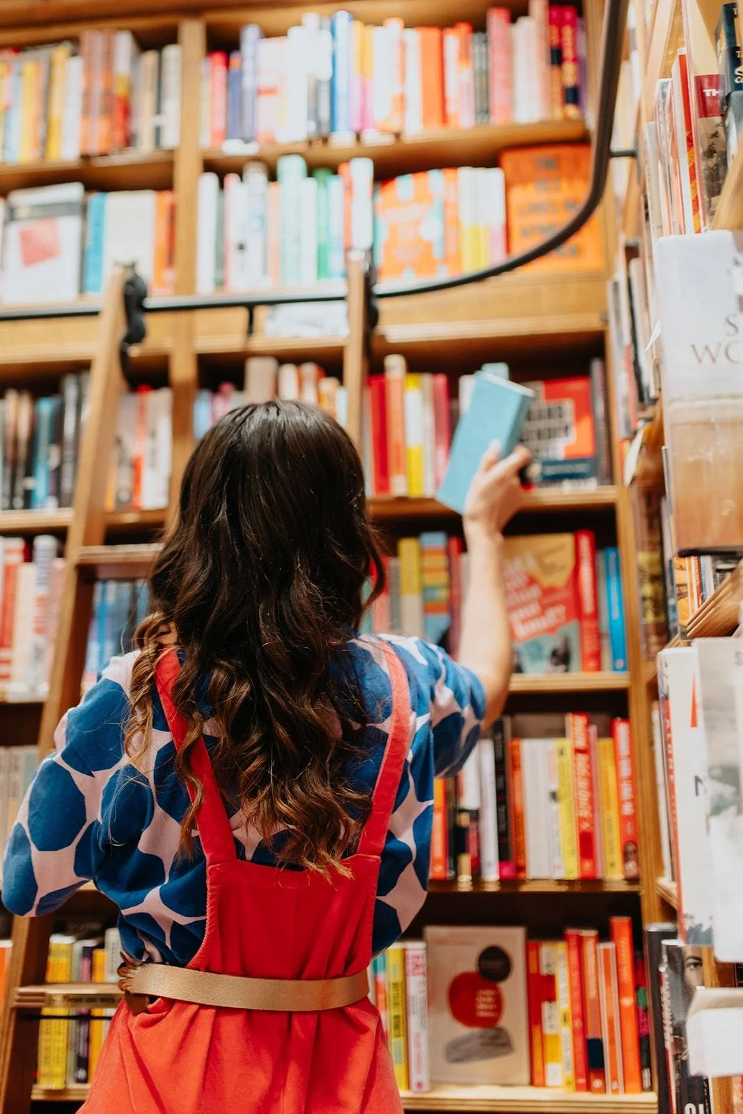 Chloe wearing a blue patterned shirt and red overalls browsing books on a wooden bookshelf in a bookstore.