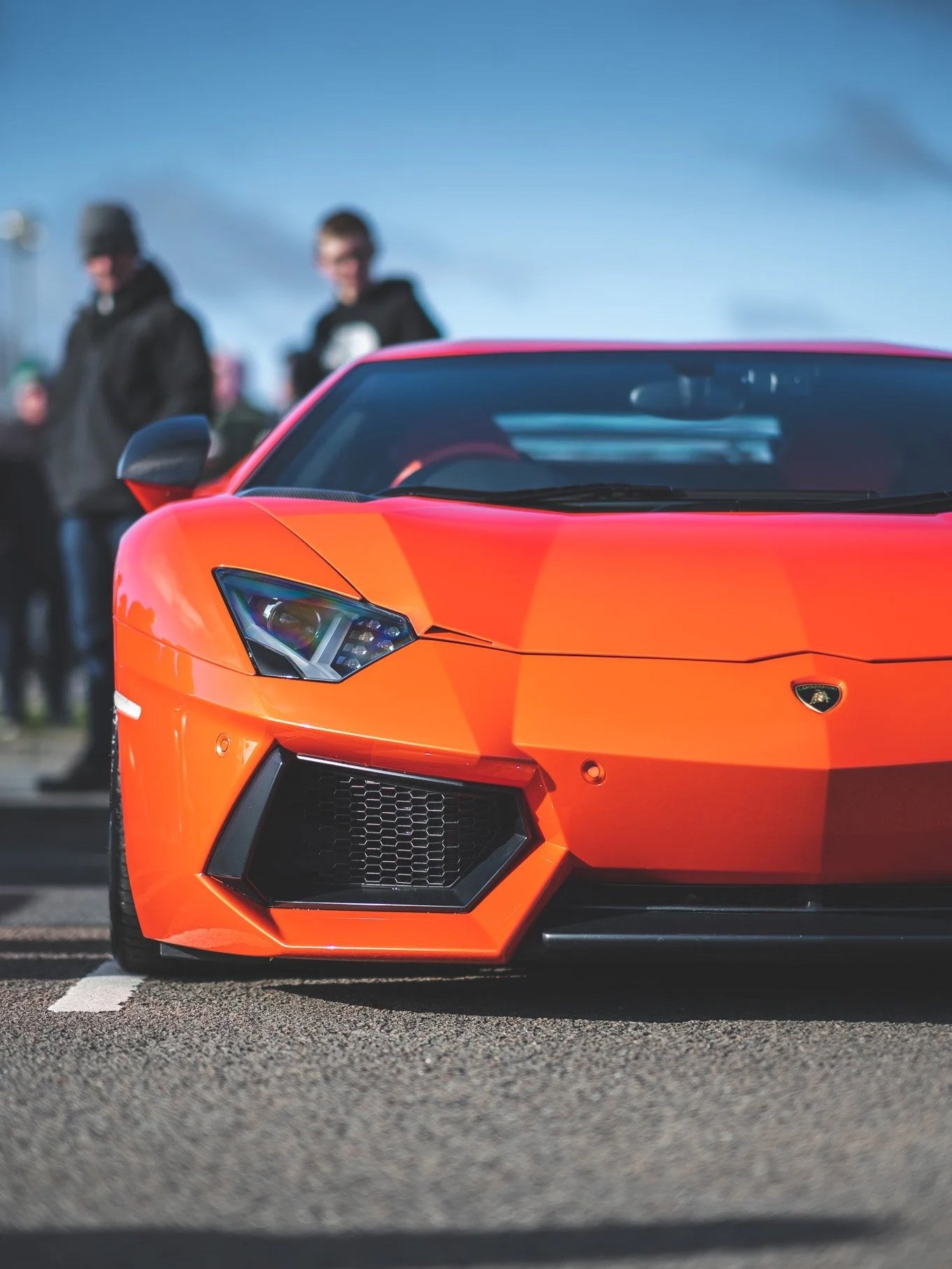 Close-up of an orange Lamborghini sports car parked on a road, with two blurred people standing in the background under a partly cloudy sky.