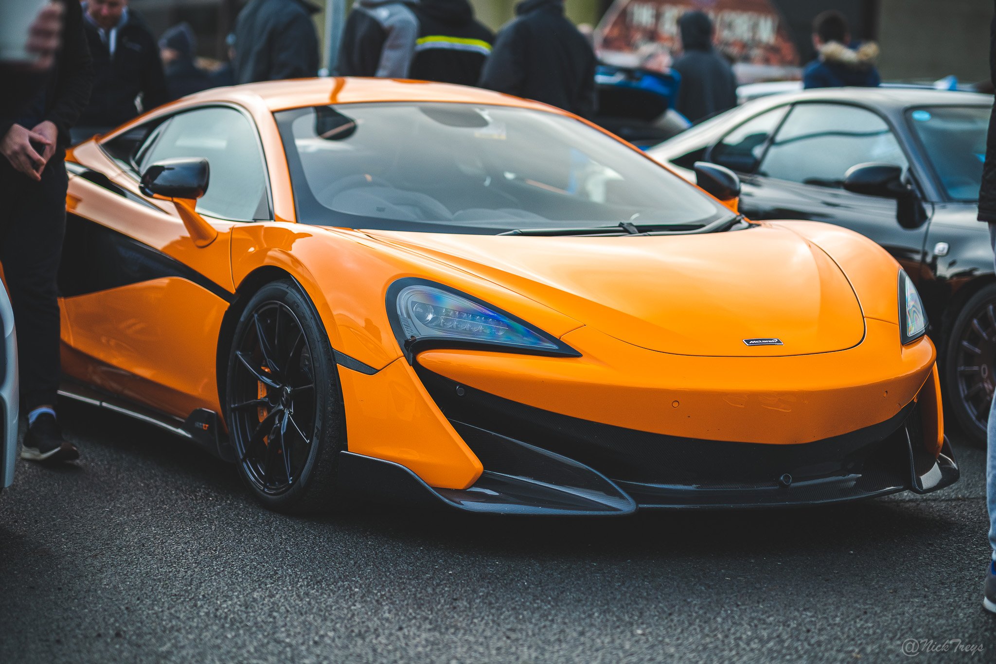 Orange McLaren sports car parked among other vehicles at an outdoor car show, with people gathered around in the background.