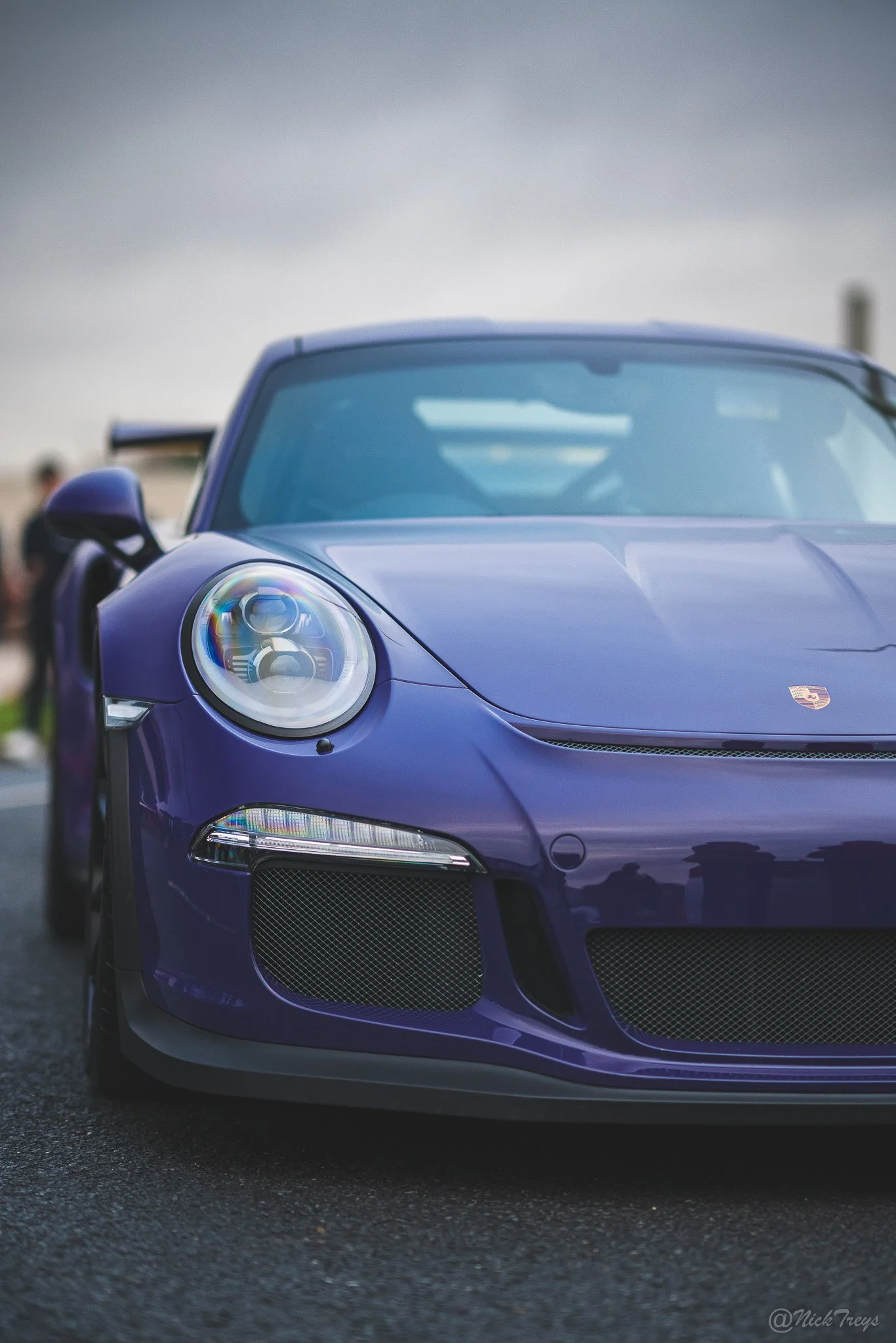 Close-up of a blue Porsche sports car showing the front left side with detailed headlight and front grille, parked on a dark asphalt surface under an overcast sky.