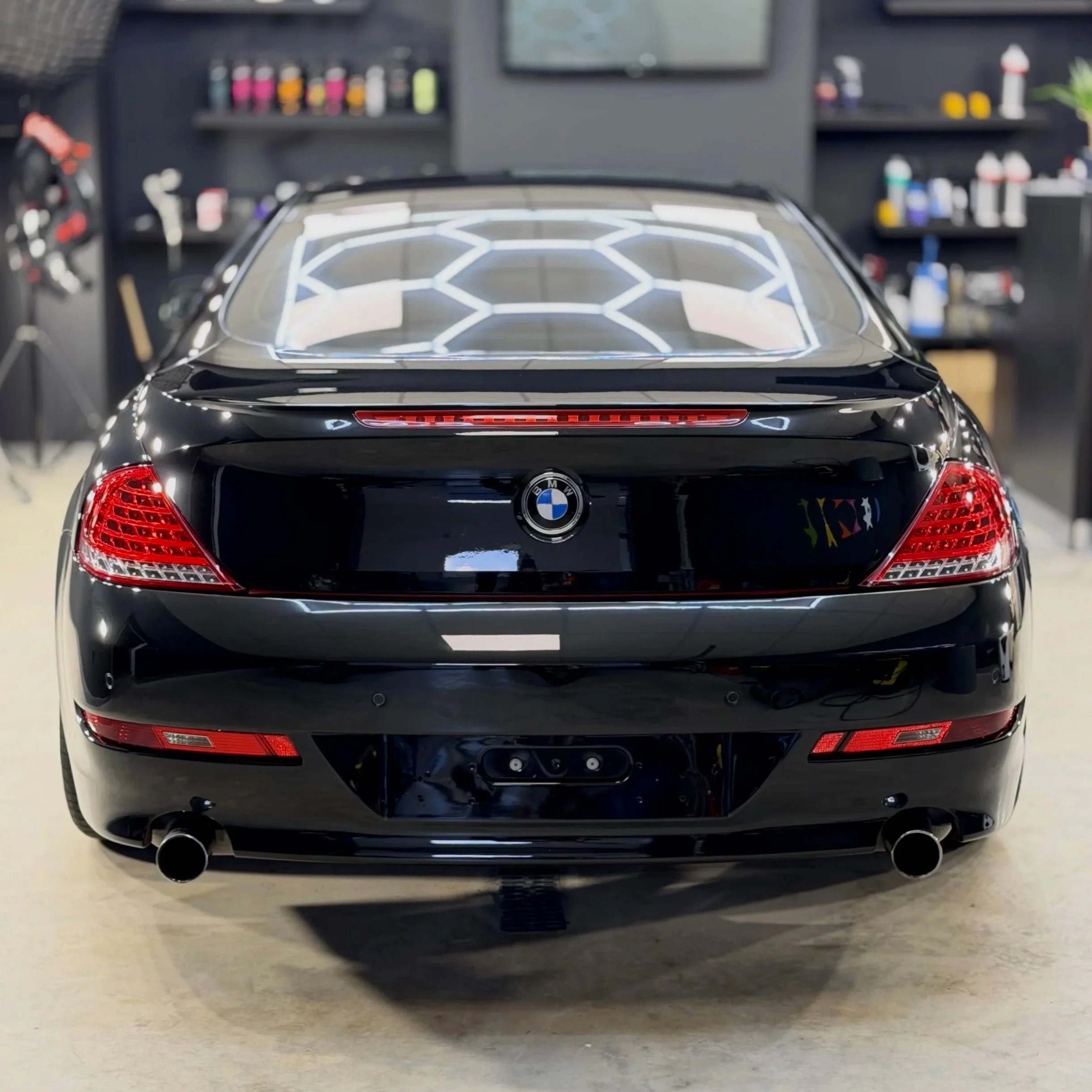 Rear view of a black BMW car with illuminated geometric light design on the back windshield, parked on a light-colored floor, with shelves of car detailing products in the background.